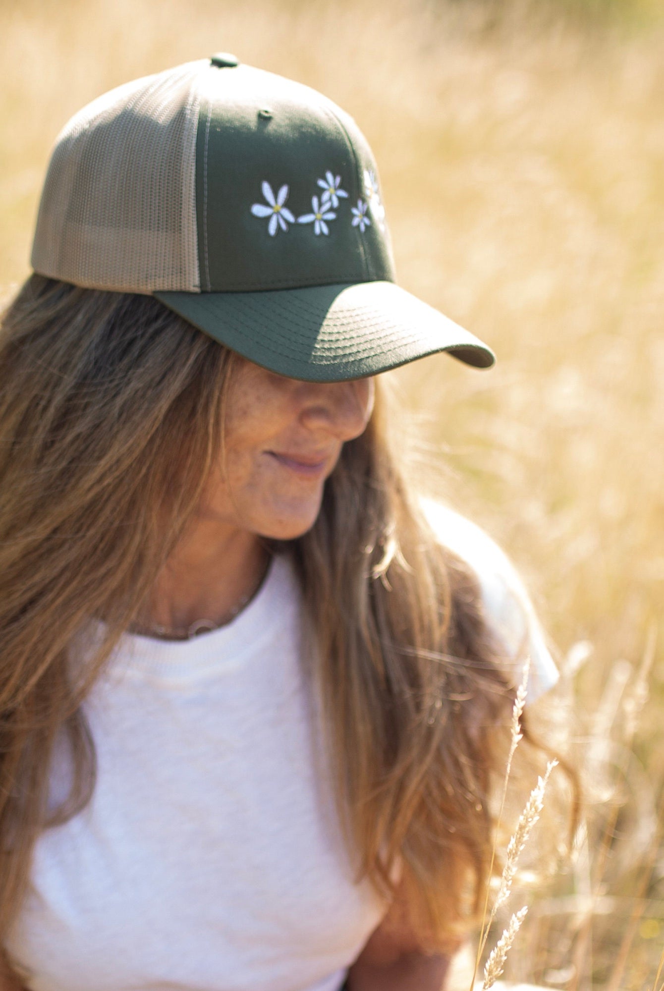 Woman with long hair is in a field of flowers. She wears a cute trucker hat that is sage green on the front panel with khaki mesh on the back and a snapback closure. Several white daisies with yellow centers are embroidered on the front of the hat.