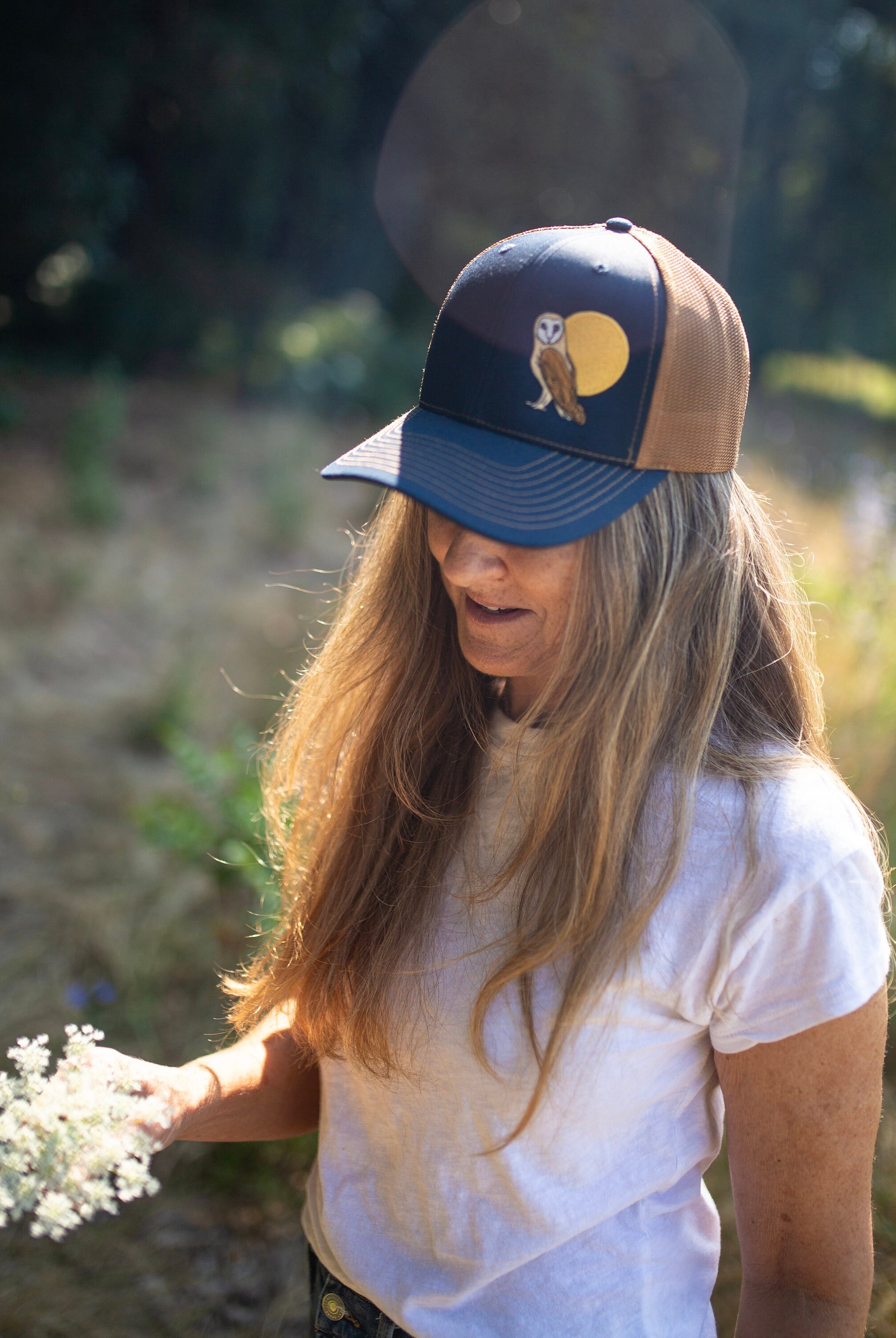 Woman with long hair is in a field of flowers. She wears a cute trucker hat that is dark blue on the front panel with khaki mesh on the back and a snapback closure. A brown and tan owl and big yellow moon is embroidered on the front of the hat.