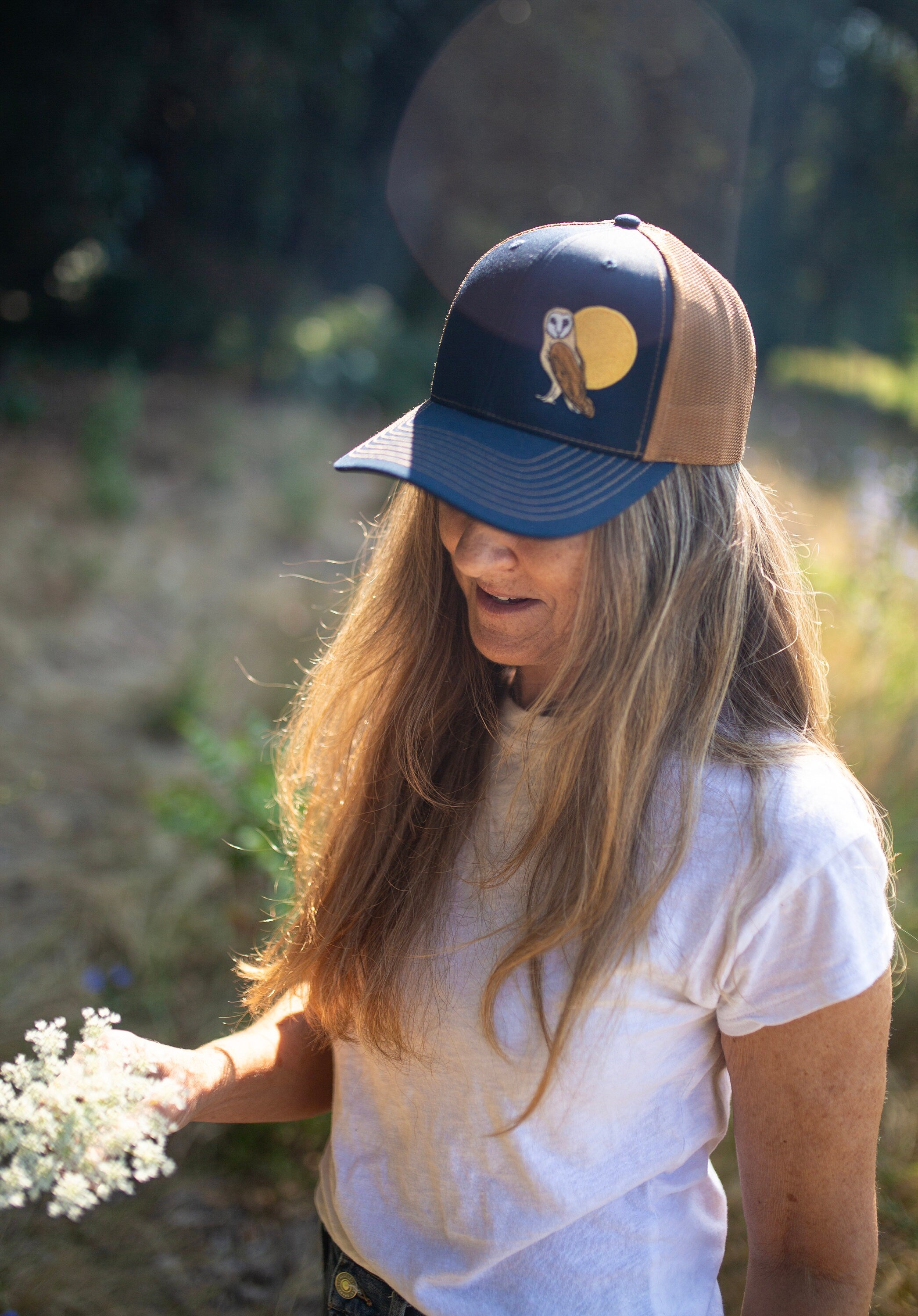 Woman with long hair is in a field of flowers. She wears a cute trucker hat that is dark blue on the front panel with khaki mesh on the back and a snapback closure. A brown and tan owl and big yellow moon is embroidered on the front of the hat.