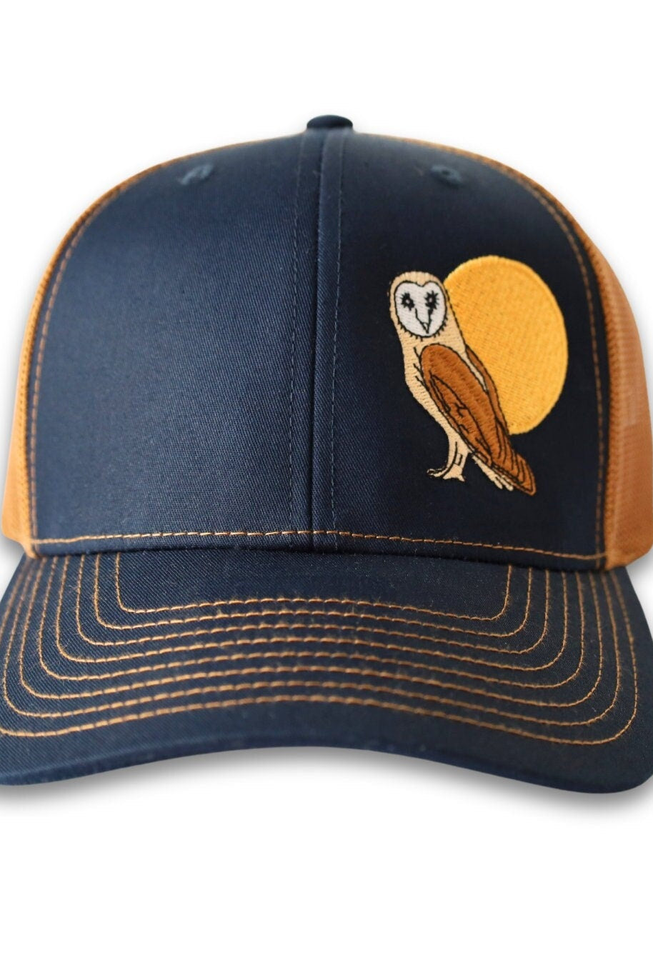 Trucker hat with a low profile. Front bill and panel is navy soft cotton fabric. A brown and tan owl and big yellow moon is embroidered on the side front. The back panel is caramel colored made of mesh with a snapback closure.
