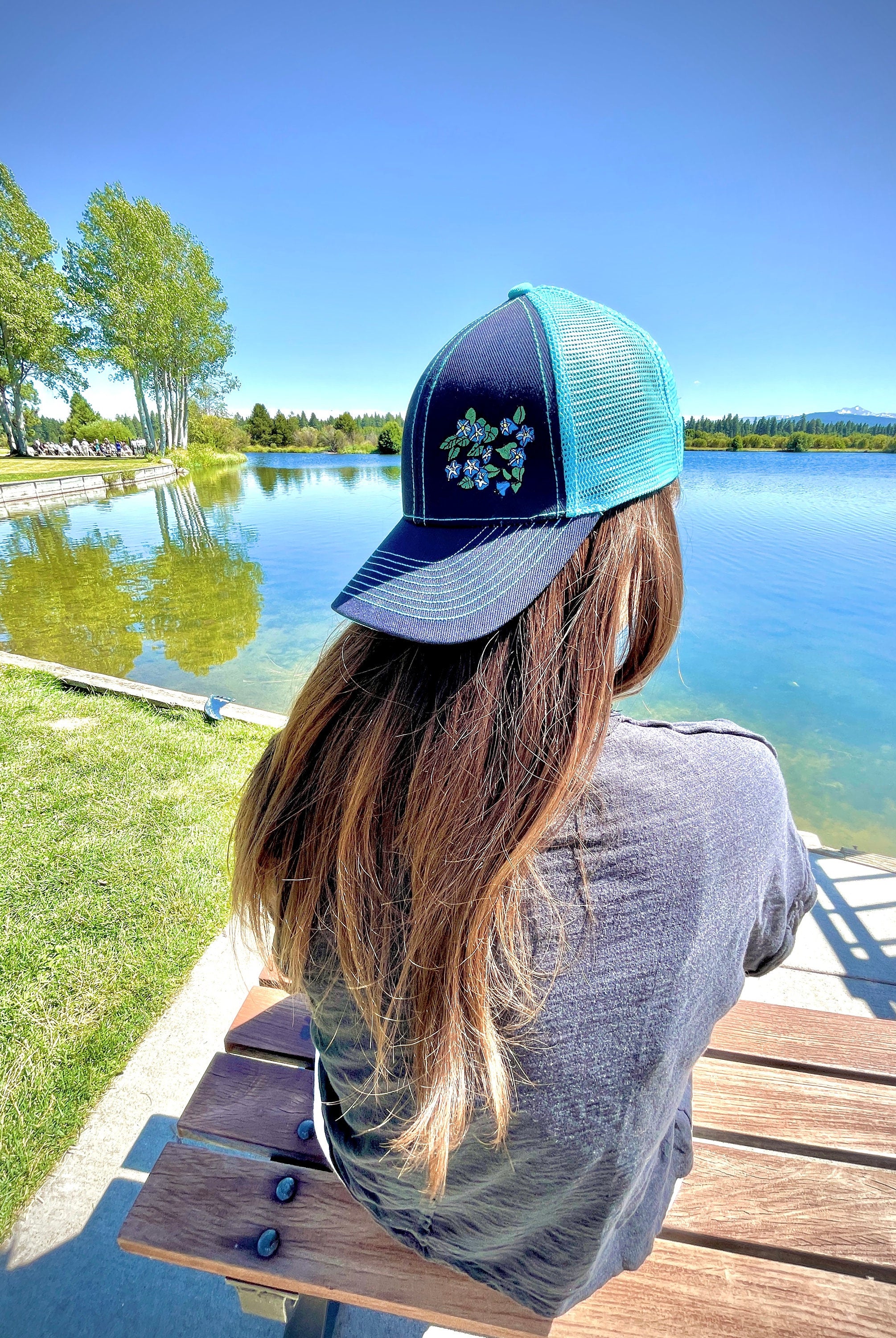 Girl with long brown hair sits facing a lake. She wears a cute trucker hat backwards that is navy blue on the front with light blue mesh on the back and a velcro closure. Morning glory flowers are embroidered on the front side of the hat.