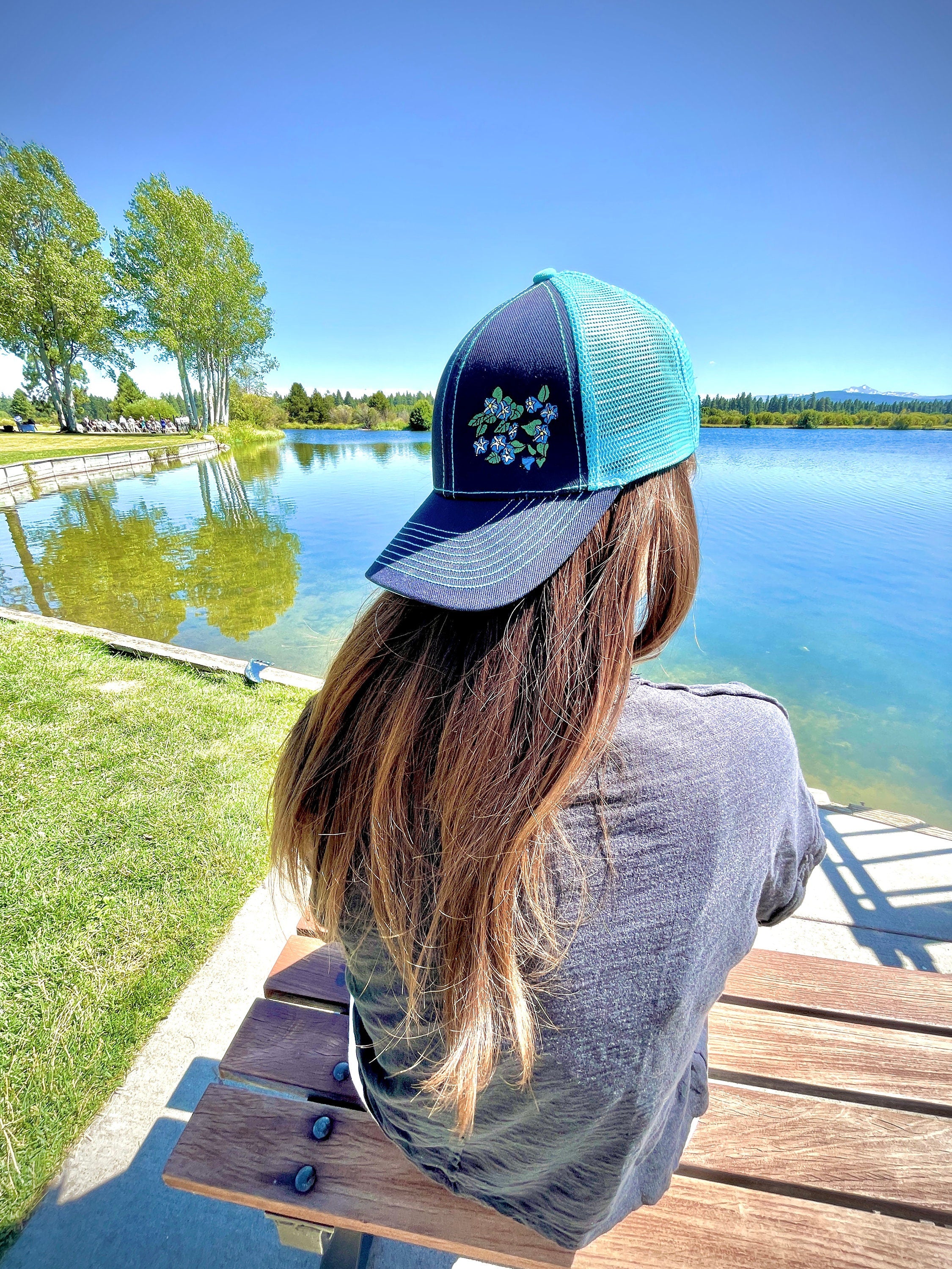 Girl with long brown hair sits facing a lake. She wears a cute trucker hat backwards that is navy blue on the front with light blue mesh on the back and a velcro closure. Morning glory flowers are embroidered on the front side of the hat.