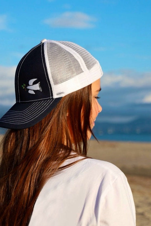 Girl with long brown hair is on the beach with the ocean. She wears a cute trucker hat that is navy blue on the front with white mesh on the back and a velcro closure. A white dove with an olive branch is embroidered on the front side of the hat.