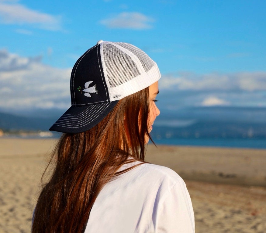 Girl with long brown hair is on the beach with the ocean. She wears a cute trucker hat that is navy blue on the front with white mesh on the back and a velcro closure. A white dove with an olive branch is embroidered on the front side of the hat.