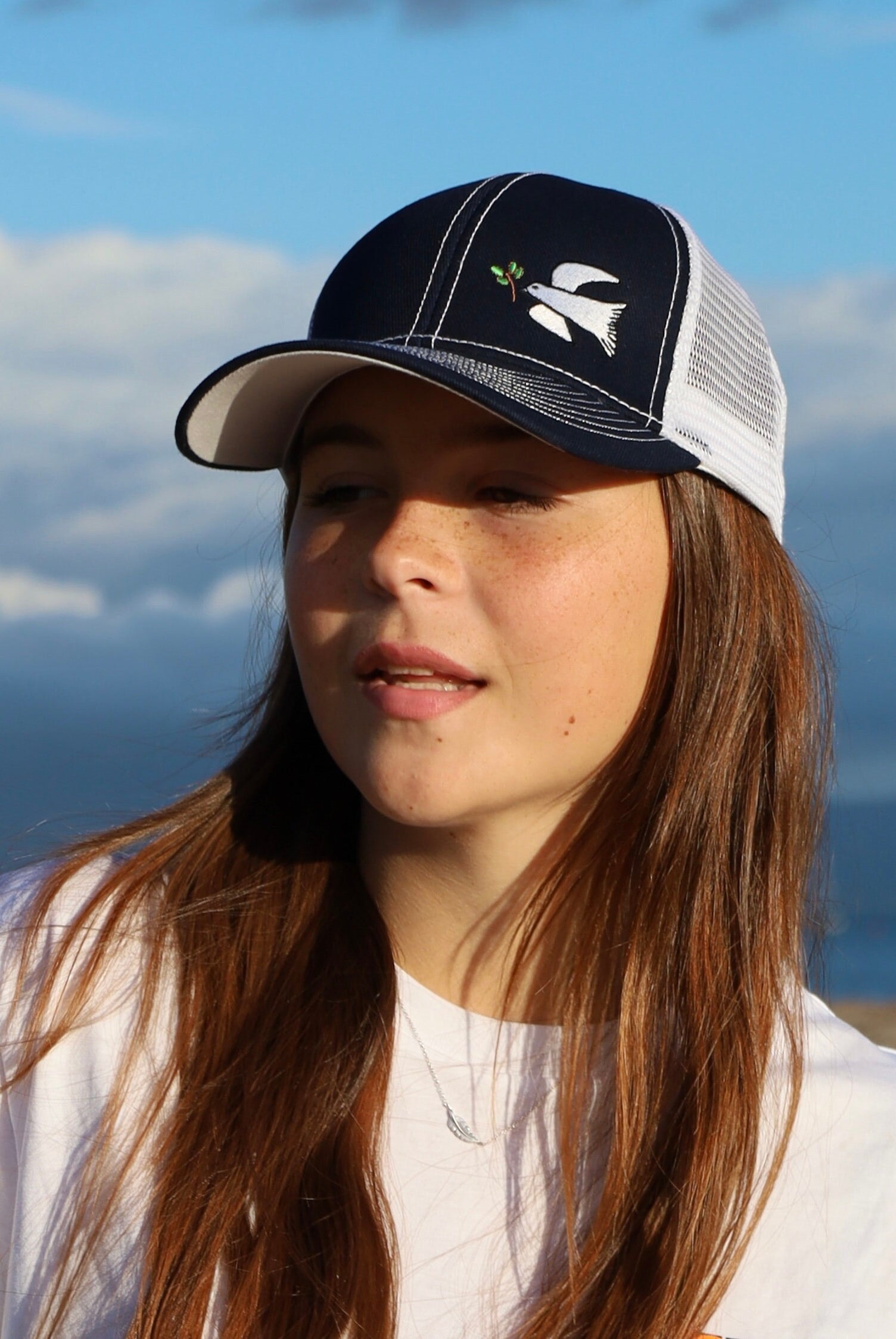 Girl with long brown hair wears a dove trucker hat that is navy blue on the front with white mesh on the back and a velcro closure. A white dove with an olive branch is embroidered on the front side of the hat.