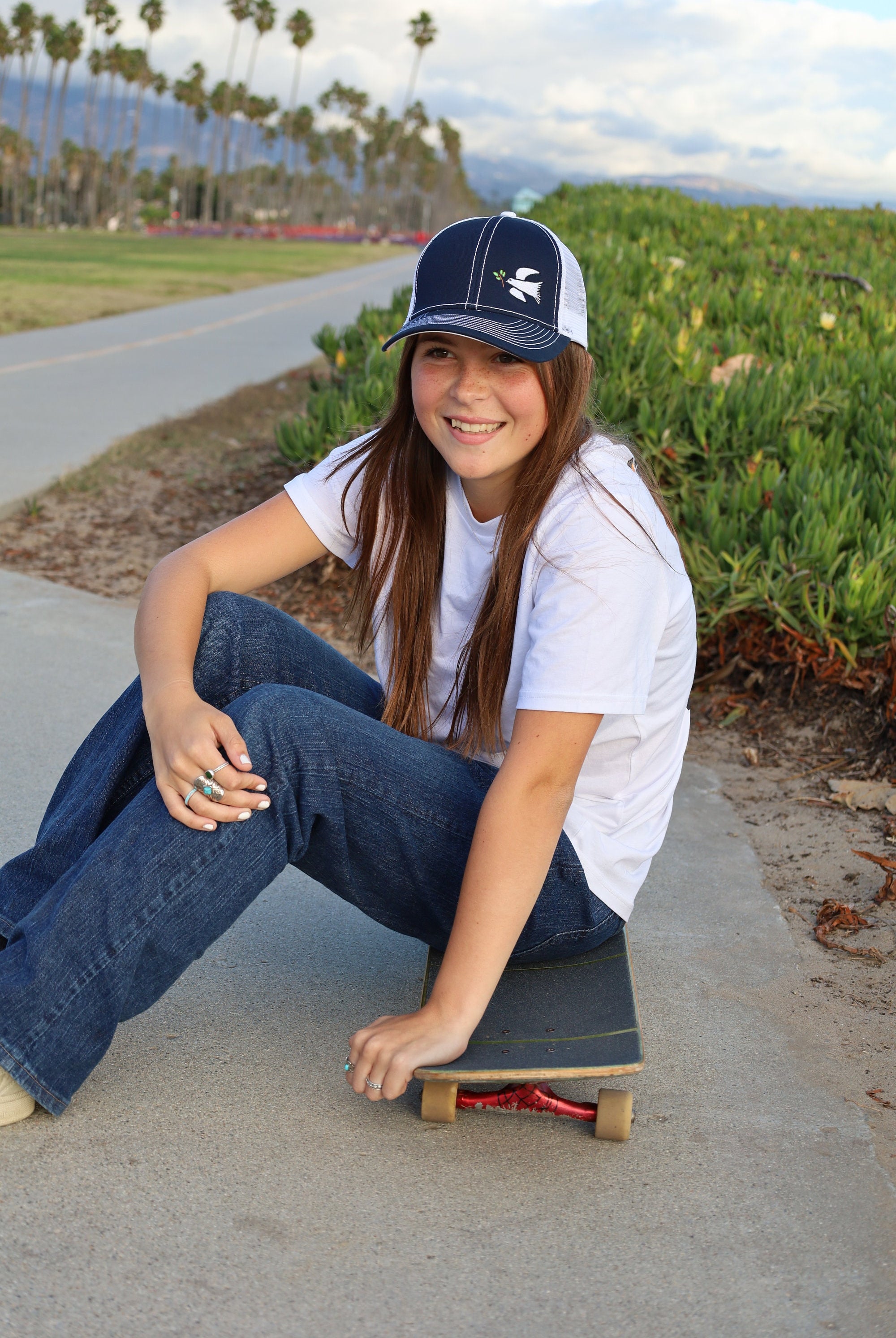 Cali skater girl with long brown hair sits on a skateboard wearing a dove trucker hat that is navy blue on the front with white mesh on the back and a velcro closure. A white dove with an olive branch is embroidered on the front side of the hat.