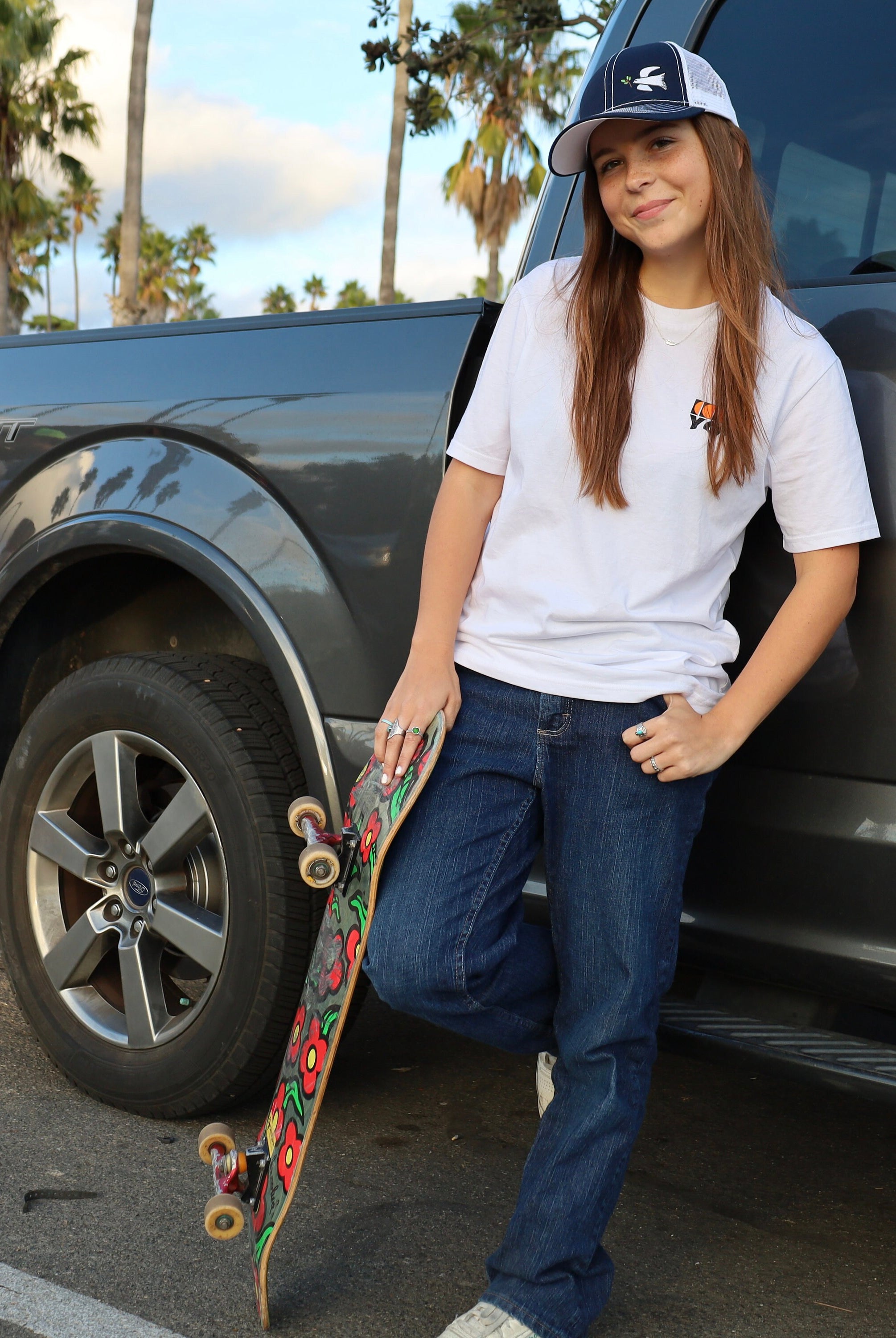 Cute skater girl with long brown hair leans on a truck wearing a dove trucker hat that is navy blue on the front with white mesh on the back and a velcro closure. A white dove with an olive branch is embroidered on the front side of the hat.