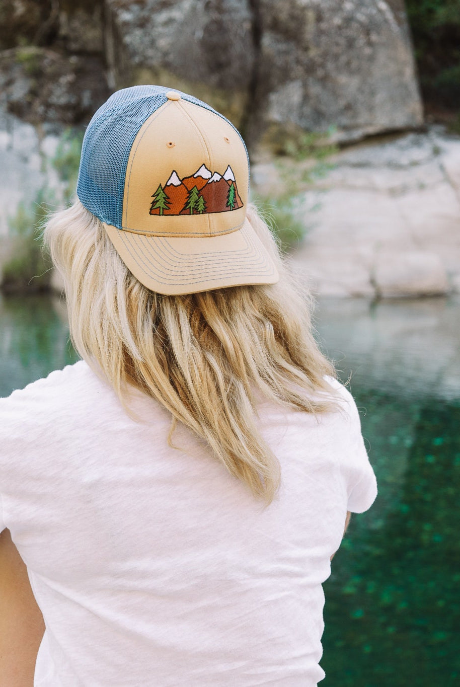 Young woman with blonde hair sits facing a river. She wears a cute trucker hat backwards that is khaki on the front panel with blue mesh on the back and a snapback closure. A mountain scene is embroidered on the front of the hat.
