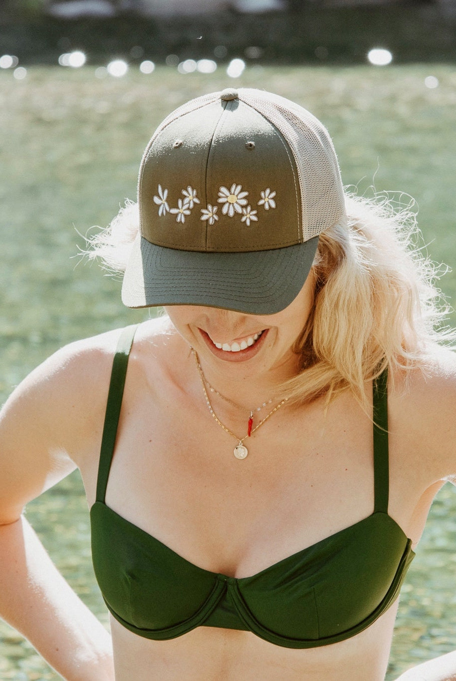 Young woman with blonde hair stands in a river. She wears a cute trucker hat that is sage green on the front panel with khaki mesh on the back and a snapback closure. Several white daisies with yellow centers are embroidered on the front of the hat.