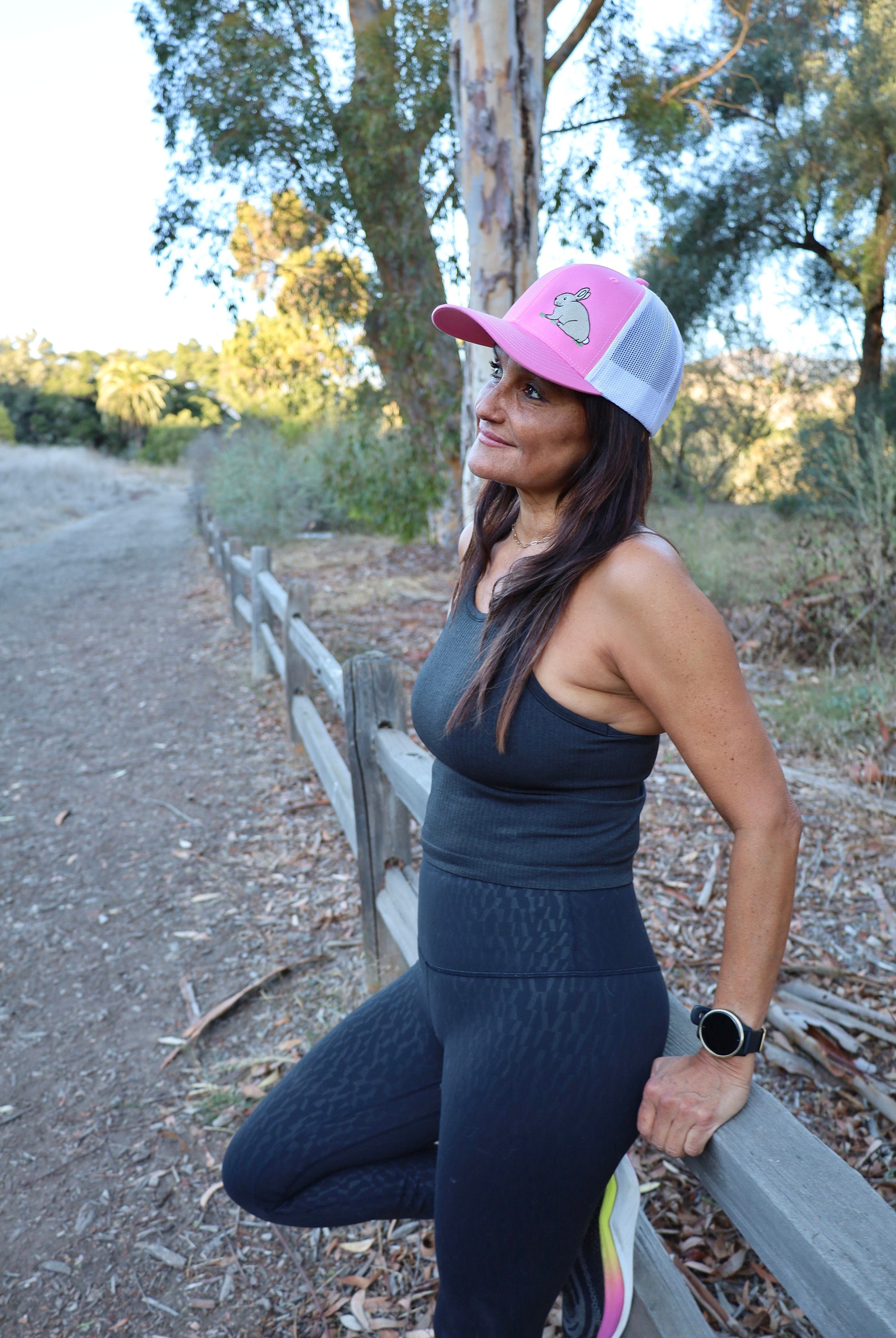 A woman with long dark hair runs on a nature trail. She is wearing a cute trucker hat that is pink on the front panel with white mesh on the back and a snapback closure. A cute tan bunny in green grass is embroidered on the front of the hat.