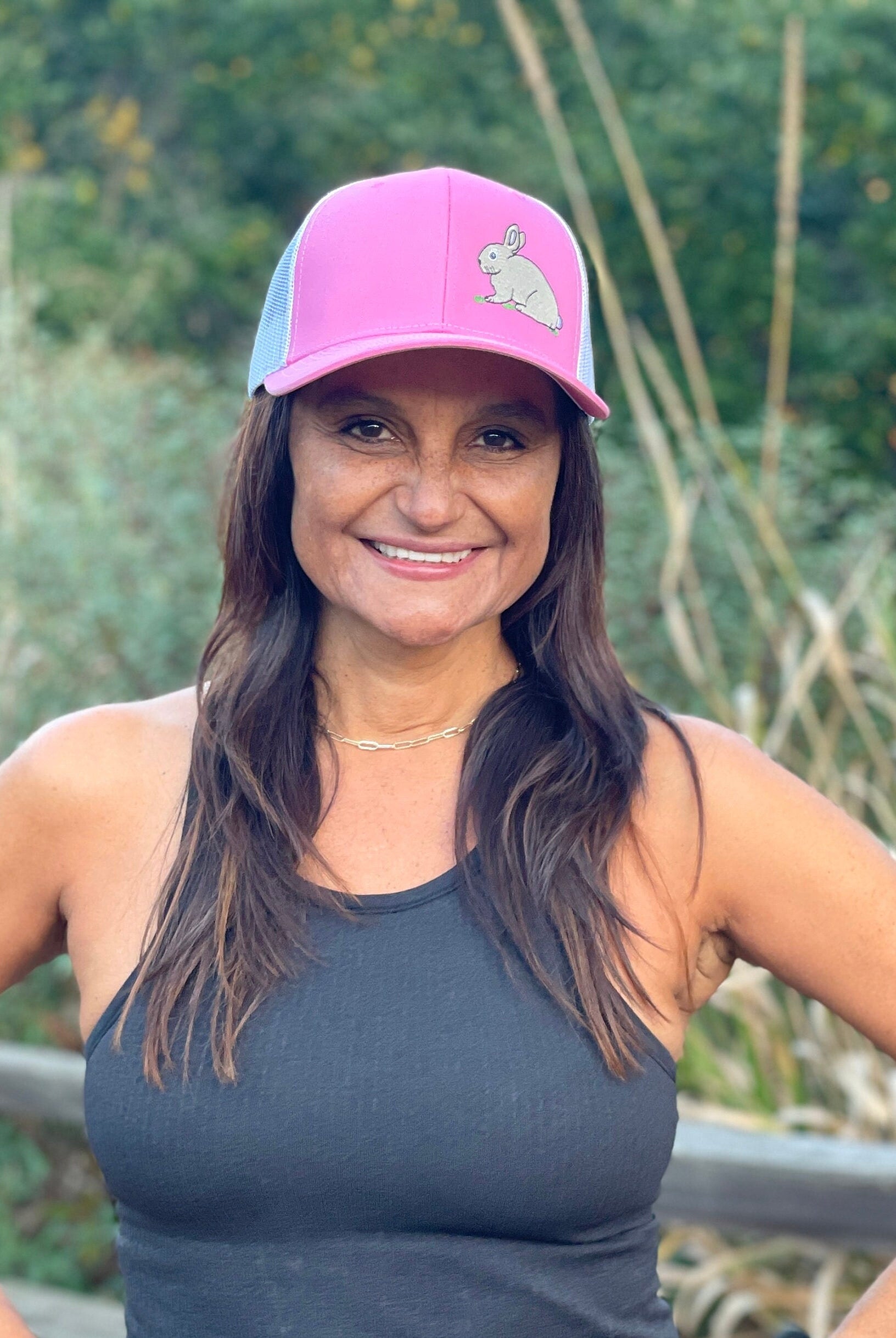 A woman with long dark hair is on a nature trail. She smiles wearing a cute trucker hat that is pink on the front panel with white mesh on the back and a snapback closure. A cute tan bunny in green grass is embroidered on the front of the hat.