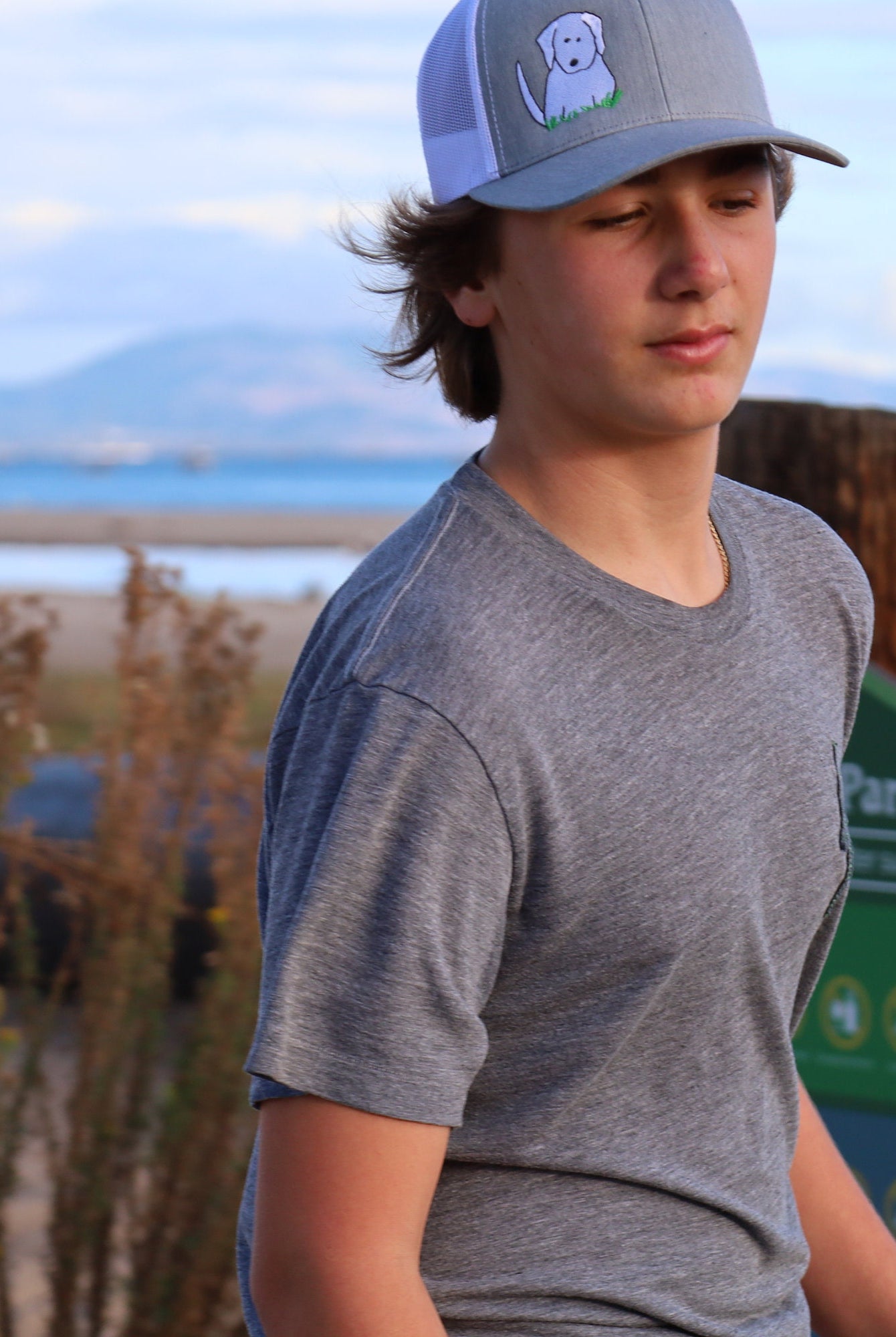 Teenage boy with brown hair stands on the beach with the ocean behind him. He wears a cute trucker hat that is grey on the front panel with white mesh on the back and a snapback closure. A cute white dog is embroidered on the front of the hat.
