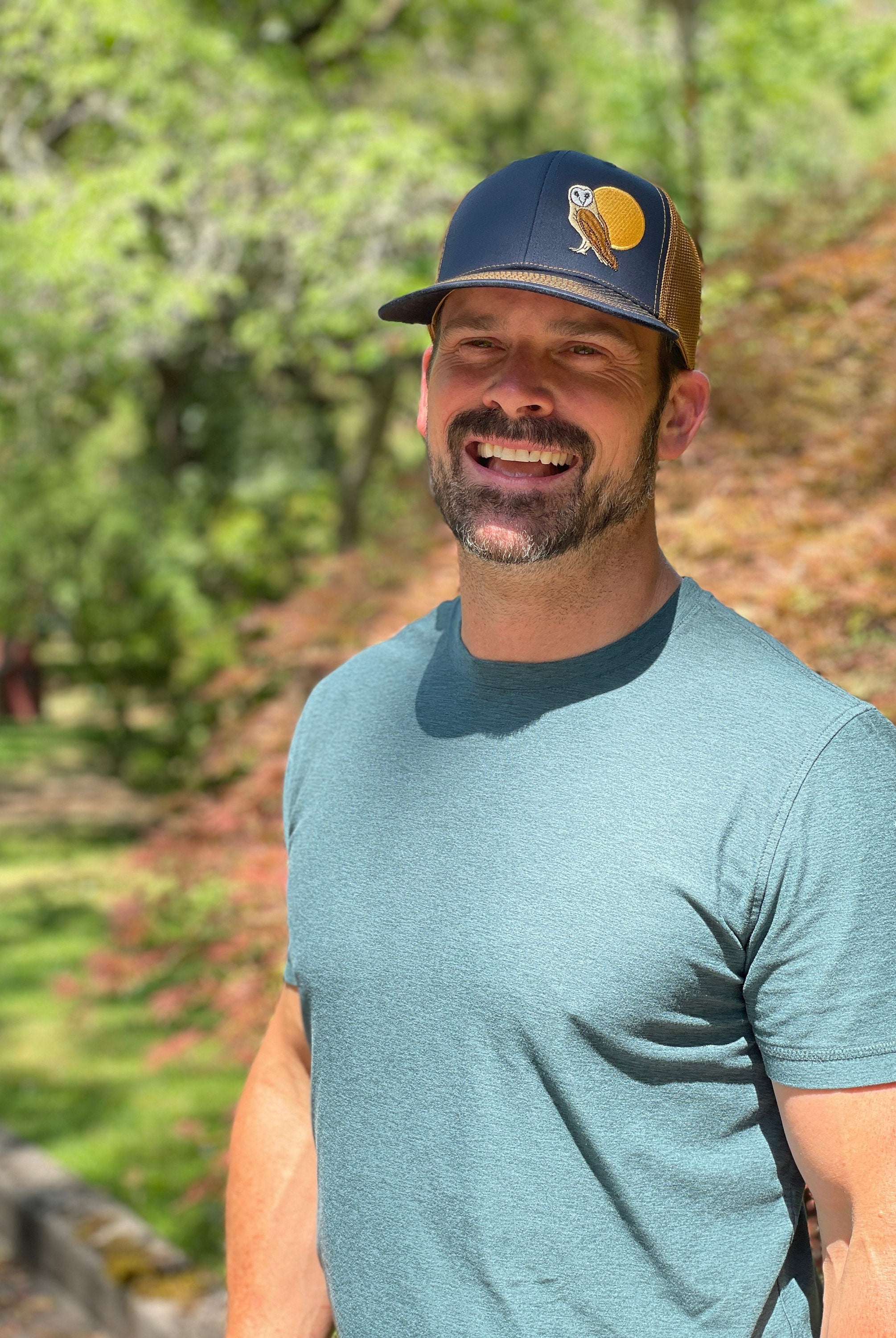 A man stands in front of trees wearing a trucker hat that is dark blue on the front panel with khaki mesh on the back and a snapback closure. A brown and tan owl and big yellow moon is embroidered on the front of the hat.
