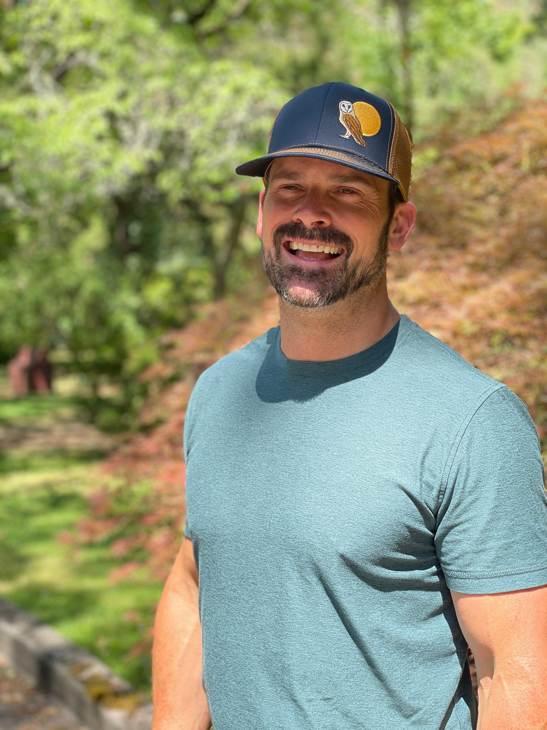 A man stands in front of trees wearing a trucker hat that is dark blue on the front panel with khaki mesh on the back and a snapback closure. A brown and tan owl and big yellow moon is embroidered on the front of the hat.