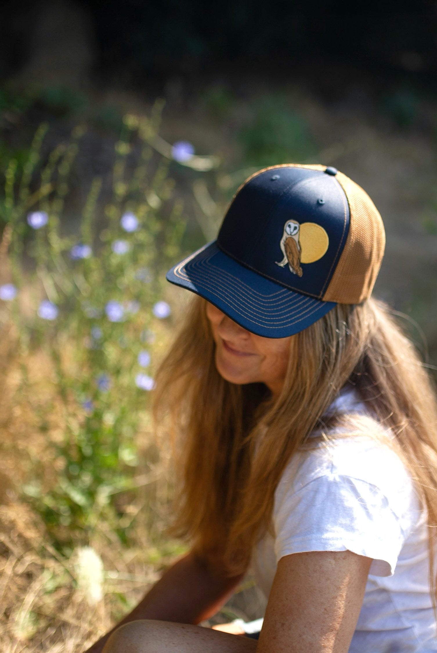 Woman with long hair is in a field of flowers. She wears a cute trucker hat that is dark blue on the front panel with khaki mesh on the back and a snapback closure. A brown and tan owl and big yellow moon is embroidered on the front of the hat.