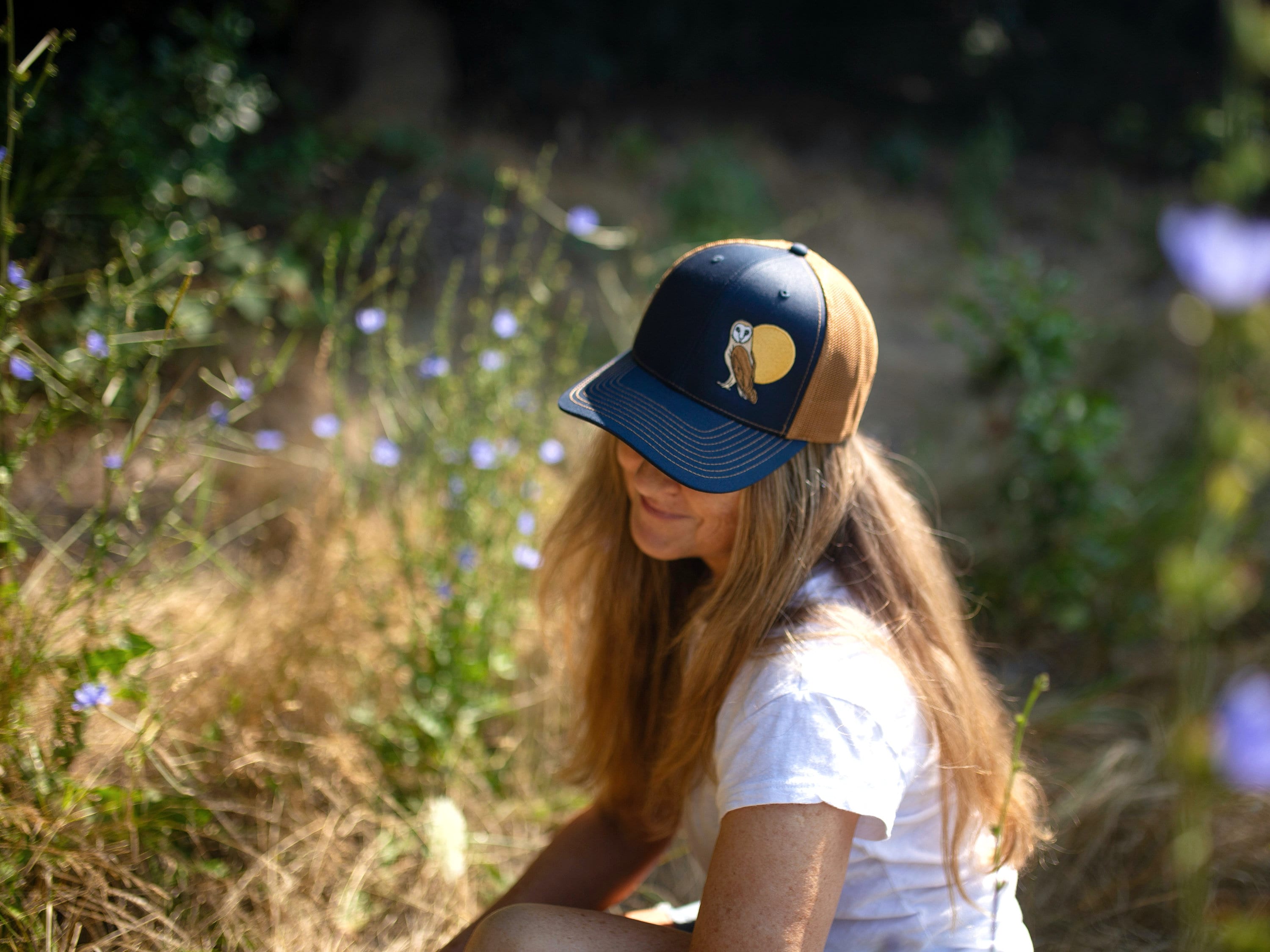 Woman with long hair is in a field of flowers. She wears a cute trucker hat that is dark blue on the front panel with khaki mesh on the back and a snapback closure. A brown and tan owl and big yellow moon is embroidered on the front of the hat.