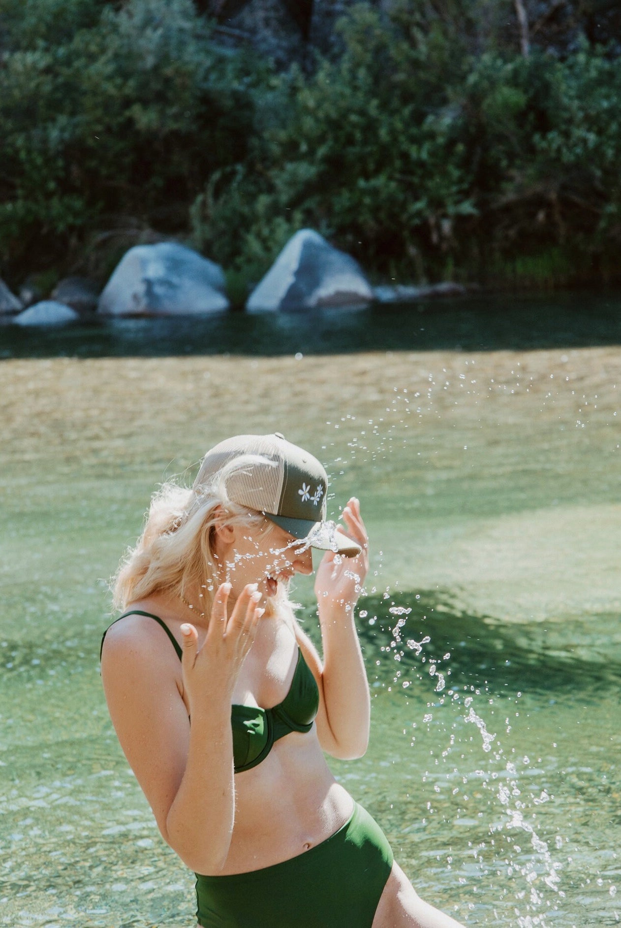 Young woman splashes water in a river. She wears a cute trucker hat that is sage green on the front panel with khaki mesh on the back and a snapback closure. Several white daisies with yellow centers are embroidered on the front of the hat.