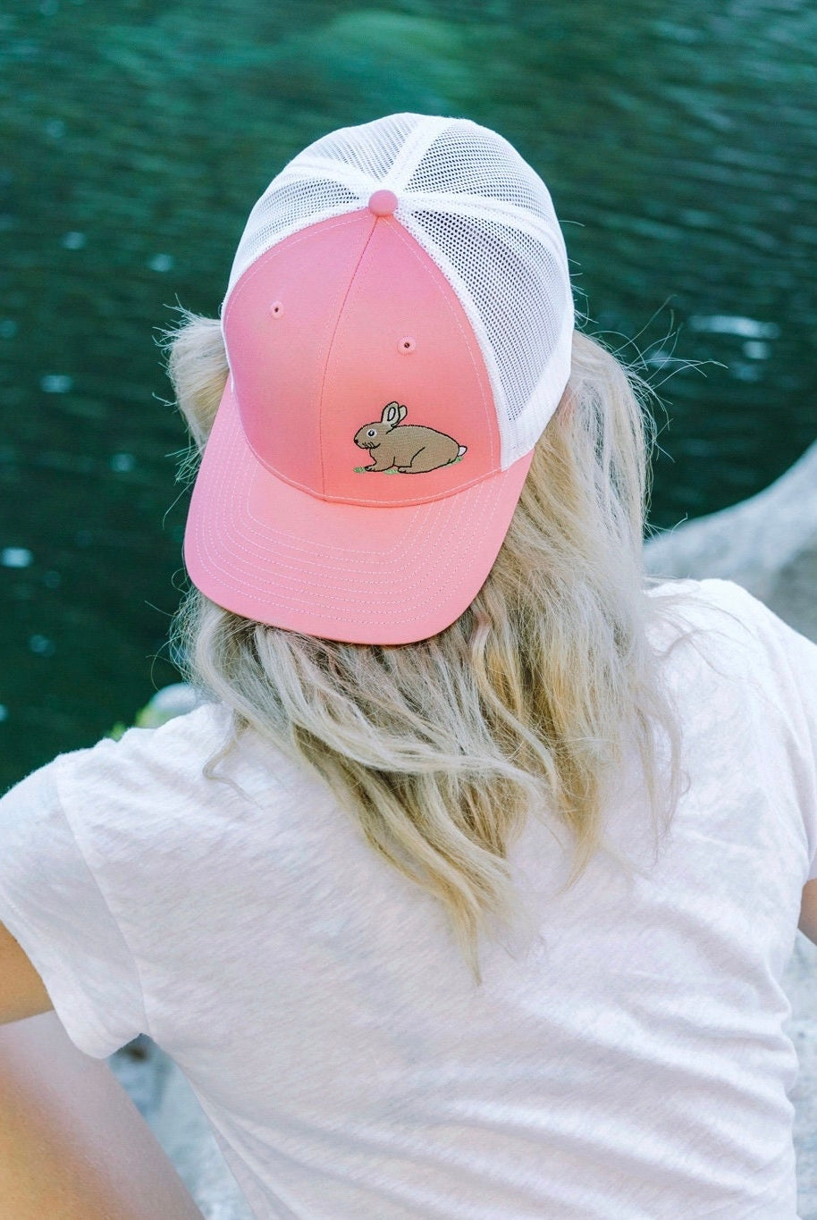 Young woman with blonde hair sits facing a river. She wears a cute trucker hat backwards that is pink on the front panel with white mesh on the back and a snapback closure. A cute tan bunny in green grass is embroidered on the front of the hat.