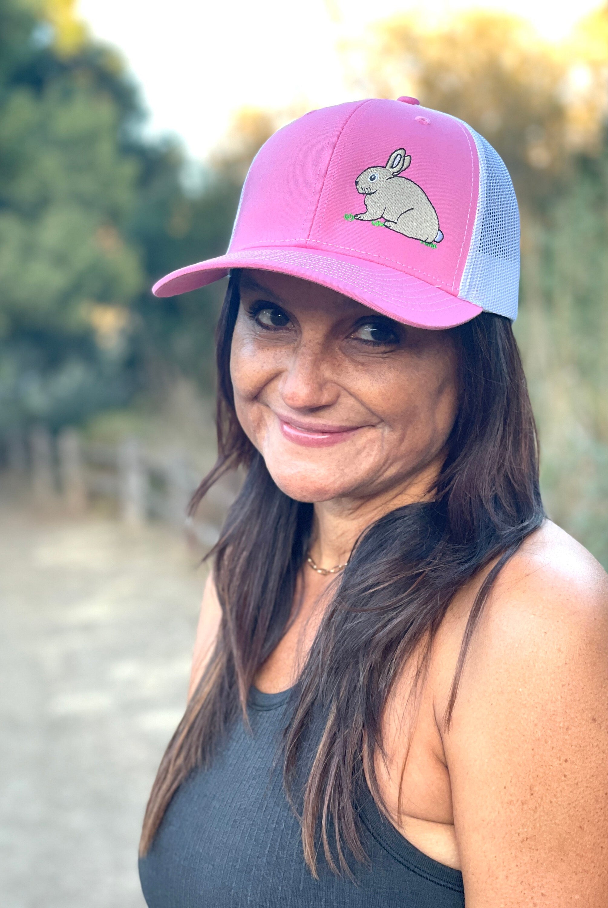 A woman with long dark hair is on a nature trail. She smiles wearing a cute trucker hat that is pink on the front panel with white mesh on the back and a snapback closure. A cute tan bunny in green grass is embroidered on the front of the hat.