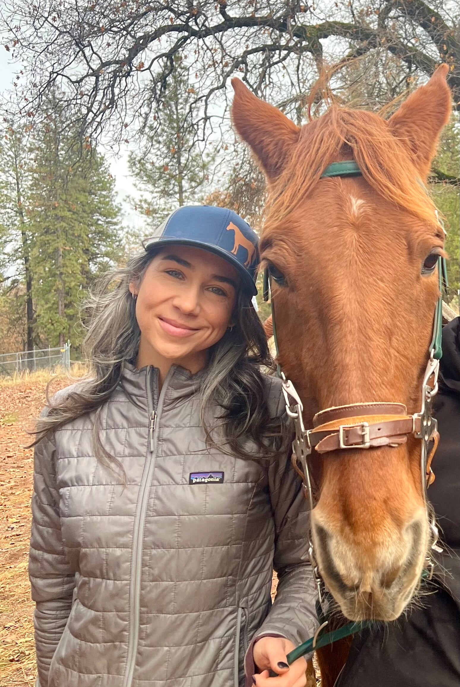 Pretty woman with long dark hair stands next to a chestnut horse and she wears a cute trucker hat that is dark blue on the front panel with white mesh on the back and a snapback closure. A chestnut horse is printed on the front side of the hat.