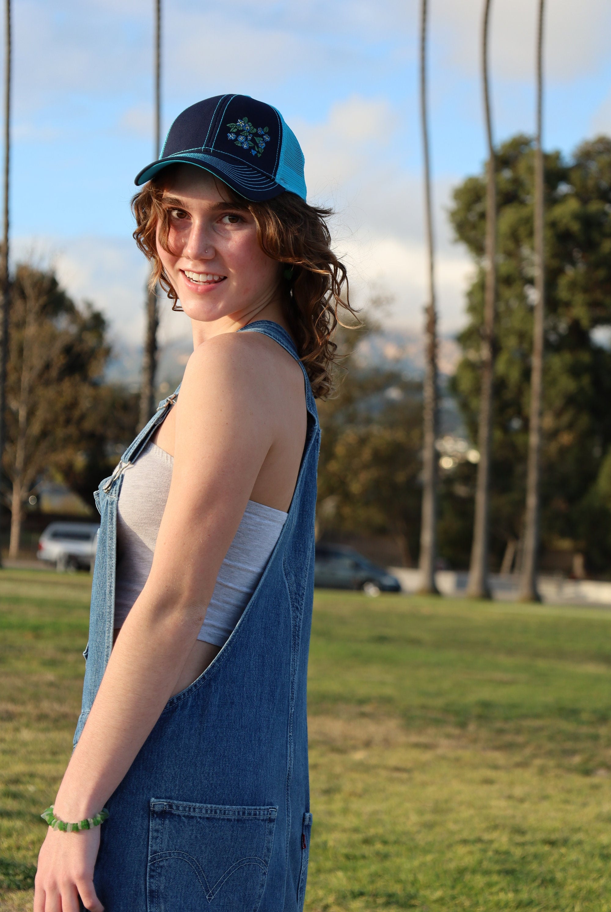 Girl with brown curly hair wears a cute trucker hat that is navy blue on the front with light blue mesh on the back and a velcro closure. Morning glory flowers are embroidered on the front side of the hat.