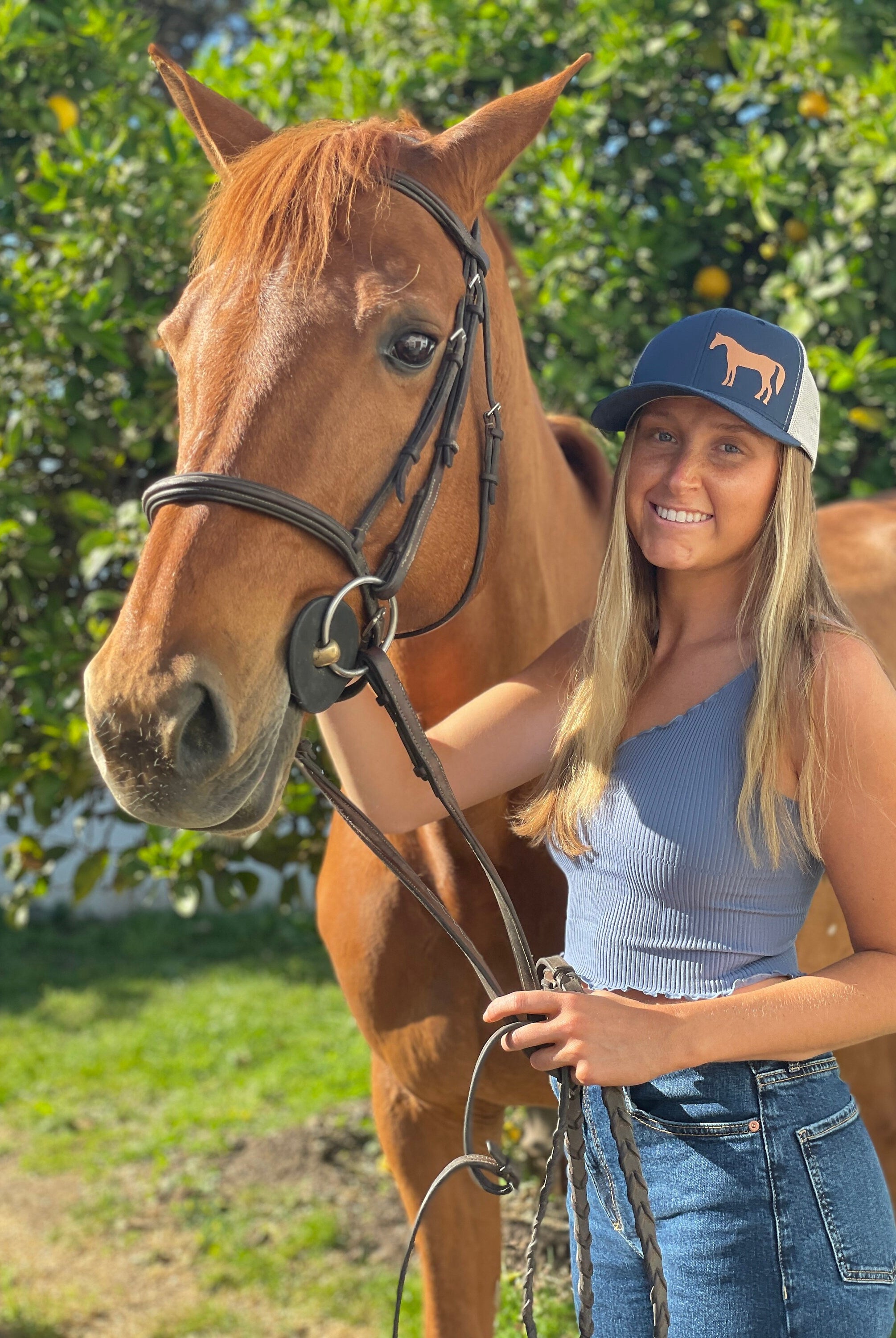 Pretty young woman with long blonde hair stands next to a chestnut horse. She wears a cute trucker hat that is dark blue on the front panel with white mesh on the back and a snapback closure. A chestnut horse is printed on the front side of the hat.