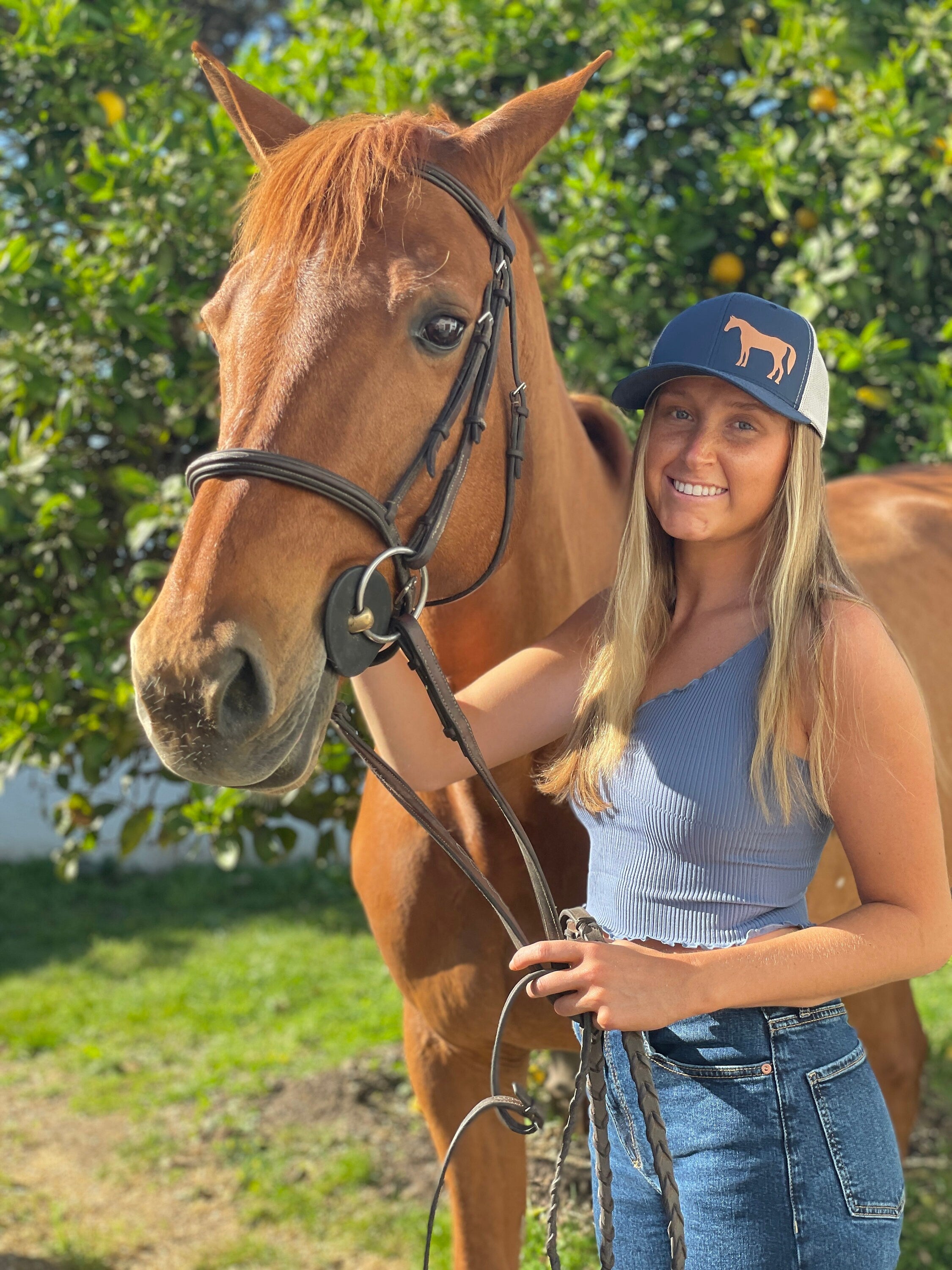 Pretty young woman with long blonde hair stands next to a chestnut horse. She wears a cute trucker hat that is dark blue on the front panel with white mesh on the back and a snapback closure. A chestnut horse is printed on the front side of the hat.
