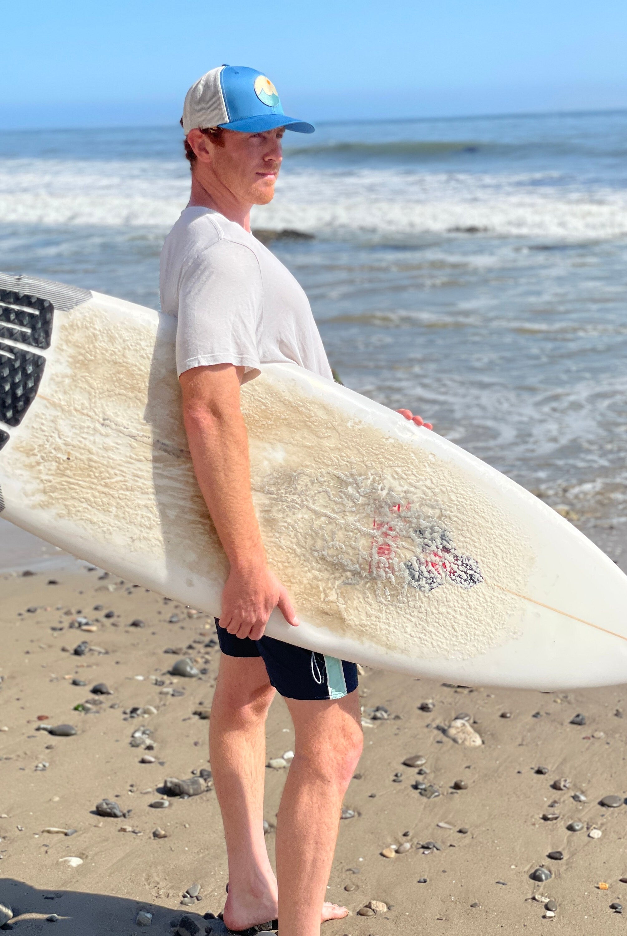 Young man is on the beach with a surf board looking at the waves. He wears a trucker hat that is blue on the front with tan mesh on the back and a snapback closure. A circle embroidered patch with ocean and sun is on the center front of the hat.