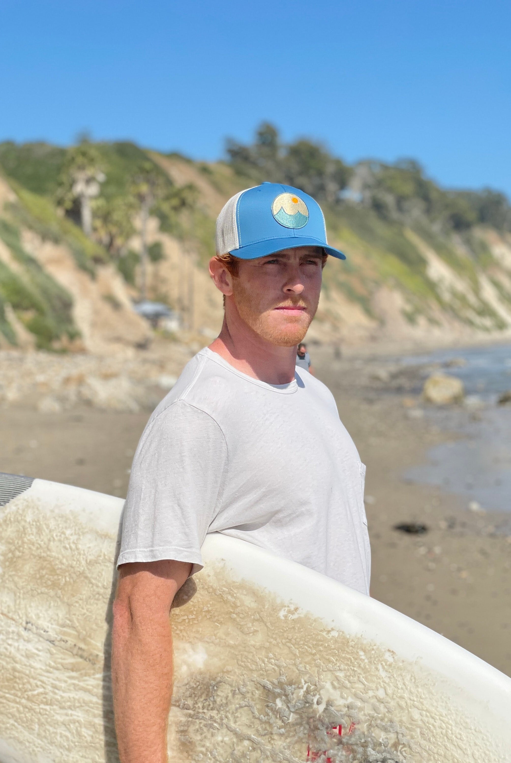 Young man is on the beach with a surf board looking at the ocean. He wears a trucker hat that is blue on the front with tan mesh on the back and a snapback closure. A circle embroidered patch with ocean and sun is on the center front of the hat.