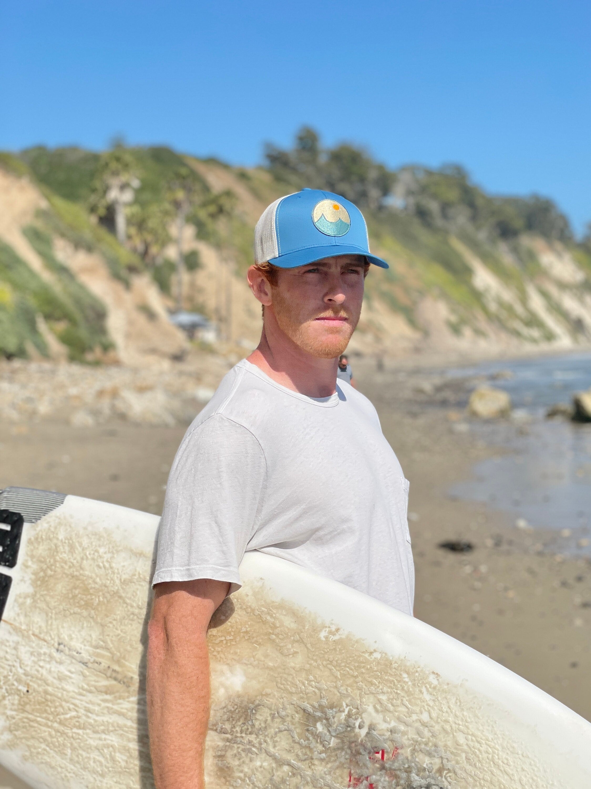 Young man is on the beach with a surf board looking at the ocean. He wears a trucker hat that is blue on the front with tan mesh on the back and a snapback closure. A circle embroidered patch with ocean and sun is on the center front of the hat.