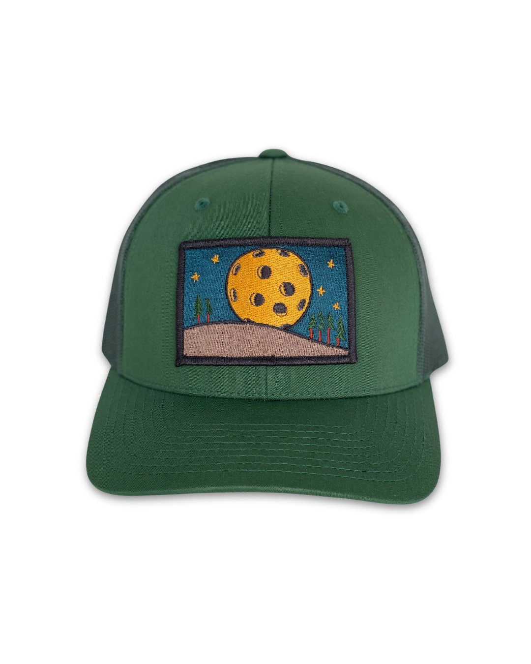 Trucker hat with a low profile. Front bill and panel is hunter green fabric. A yellow pickleball moon over a tan hill with green trees is embroidered on a patch in the center. The back panel is also green made of mesh with a snapback closure.