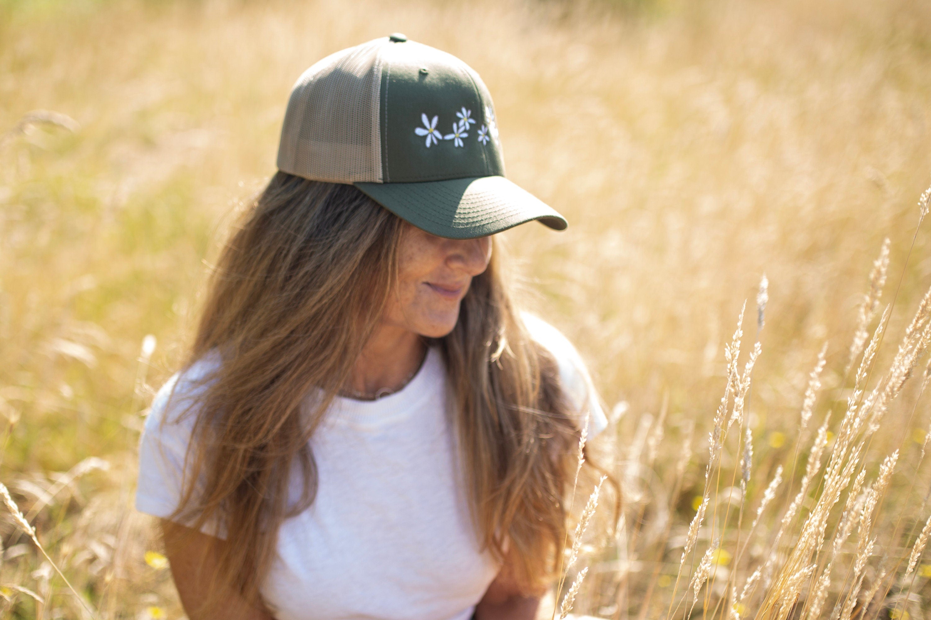 Woman with long hair is in a field of flowers. She wears a cute trucker hat that is sage green on the front panel with khaki mesh on the back and a snapback closure. Several white daisies with yellow centers are embroidered on the front of the hat.