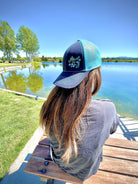 Girl with long brown hair sits facing a lake. She wears a cute trucker hat backwards that is navy blue on the front with light blue mesh on the back and a velcro closure. Morning glory flowers are embroidered on the front side of the hat.