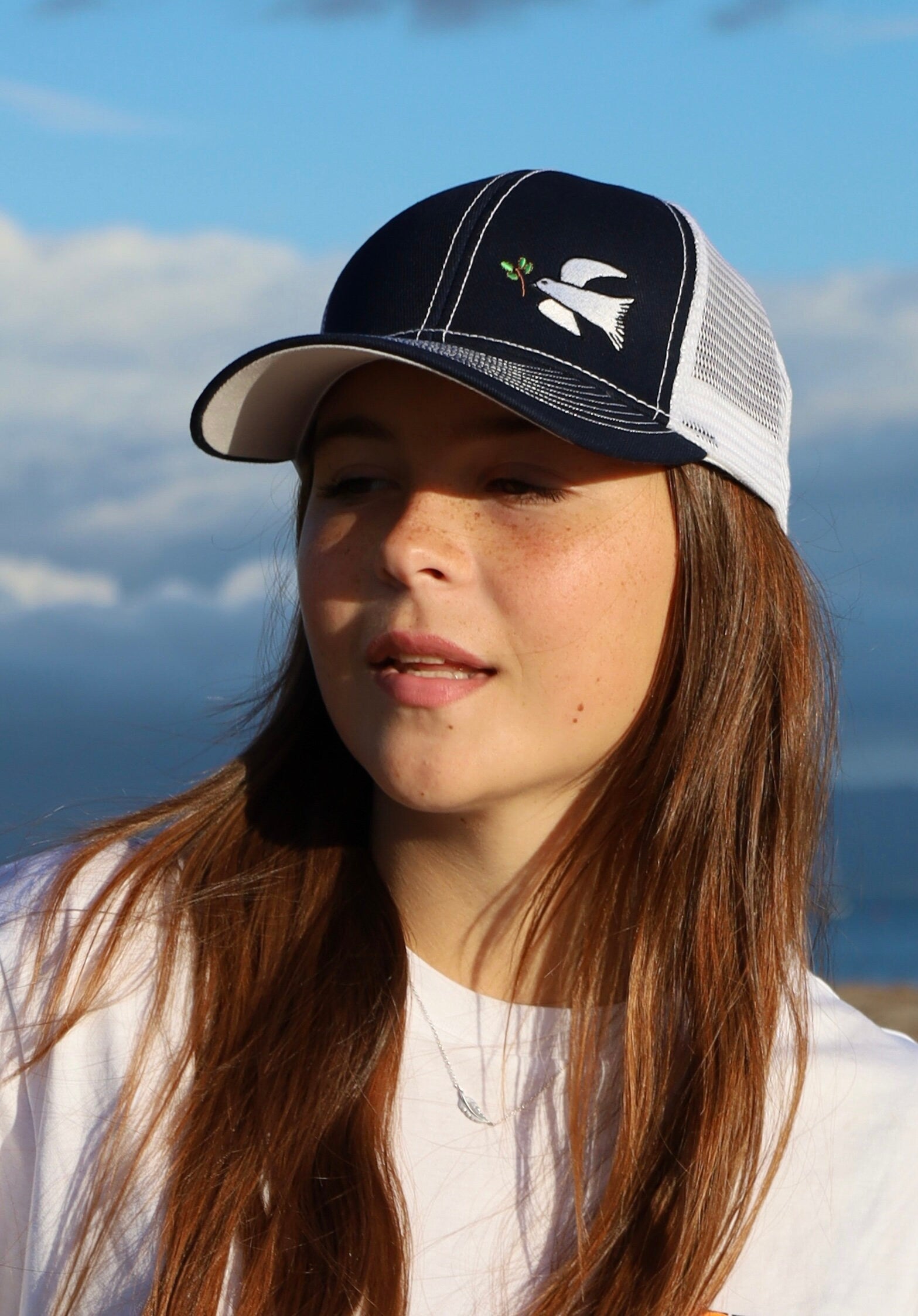 Girl with long brown hair wears a dove trucker hat that is navy blue on the front with white mesh on the back and a velcro closure. A white dove with an olive branch is embroidered on the front side of the hat.