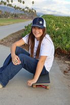Cali skater girl with long brown hair sits on a skateboard wearing a dove trucker hat that is navy blue on the front with white mesh on the back and a velcro closure. A white dove with an olive branch is embroidered on the front side of the hat.
