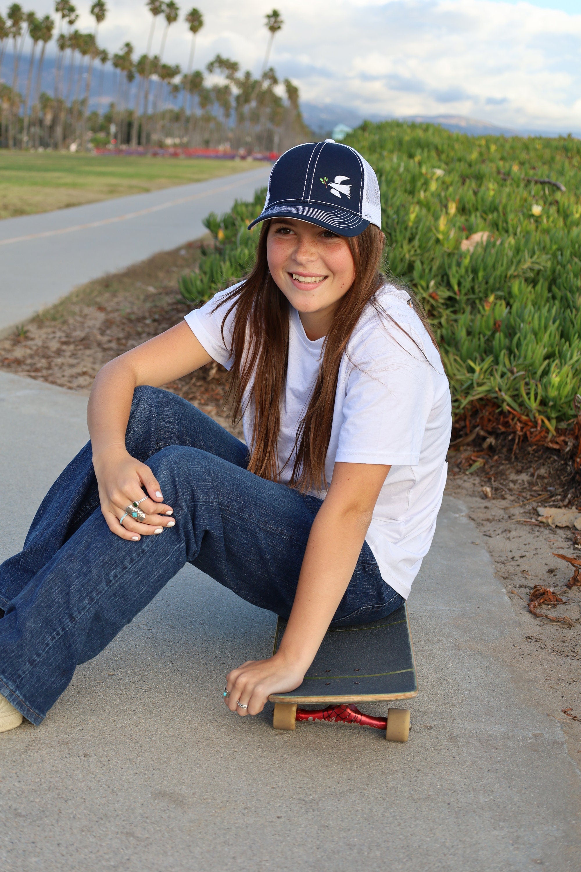 Cali skater girl with long brown hair sits on a skateboard wearing a dove trucker hat that is navy blue on the front with white mesh on the back and a velcro closure. A white dove with an olive branch is embroidered on the front side of the hat.