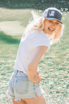 Young woman with blonde hair laughs by a river. She wears a cute trucker hat that is grey on the front panel with white mesh on the back and a snapback closure. A cute white dog is embroidered on the front of the hat.