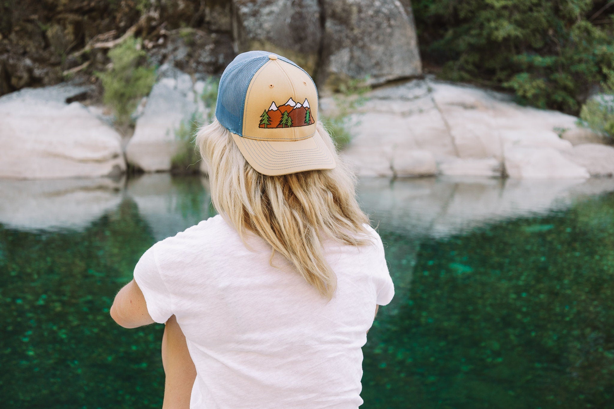 Young woman with blonde hair sits facing a river. She wears a cute trucker hat backwards that is khaki on the front panel with blue mesh on the back and a snapback closure. A mountain scene is embroidered on the front of the hat.