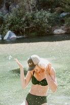 Young woman splashes water in a river. She wears a cute trucker hat that is sage green on the front panel with khaki mesh on the back and a snapback closure. Several white daisies with yellow centers are embroidered on the front of the hat.