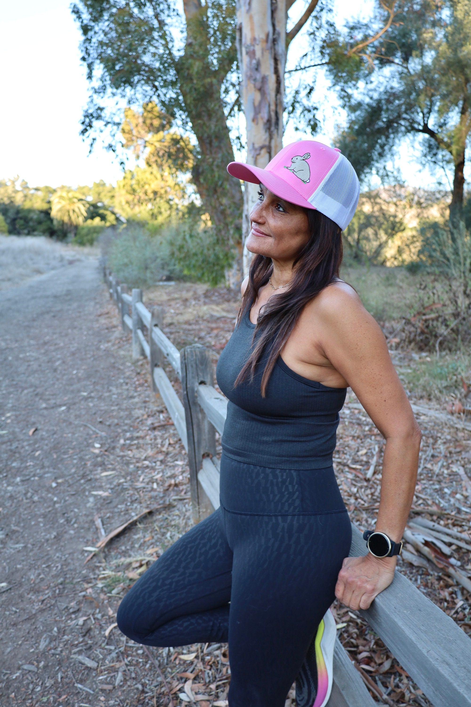A woman with long dark hair runs on a nature trail. She is wearing a cute trucker hat that is pink on the front panel with white mesh on the back and a snapback closure. A cute tan bunny in green grass is embroidered on the front of the hat.