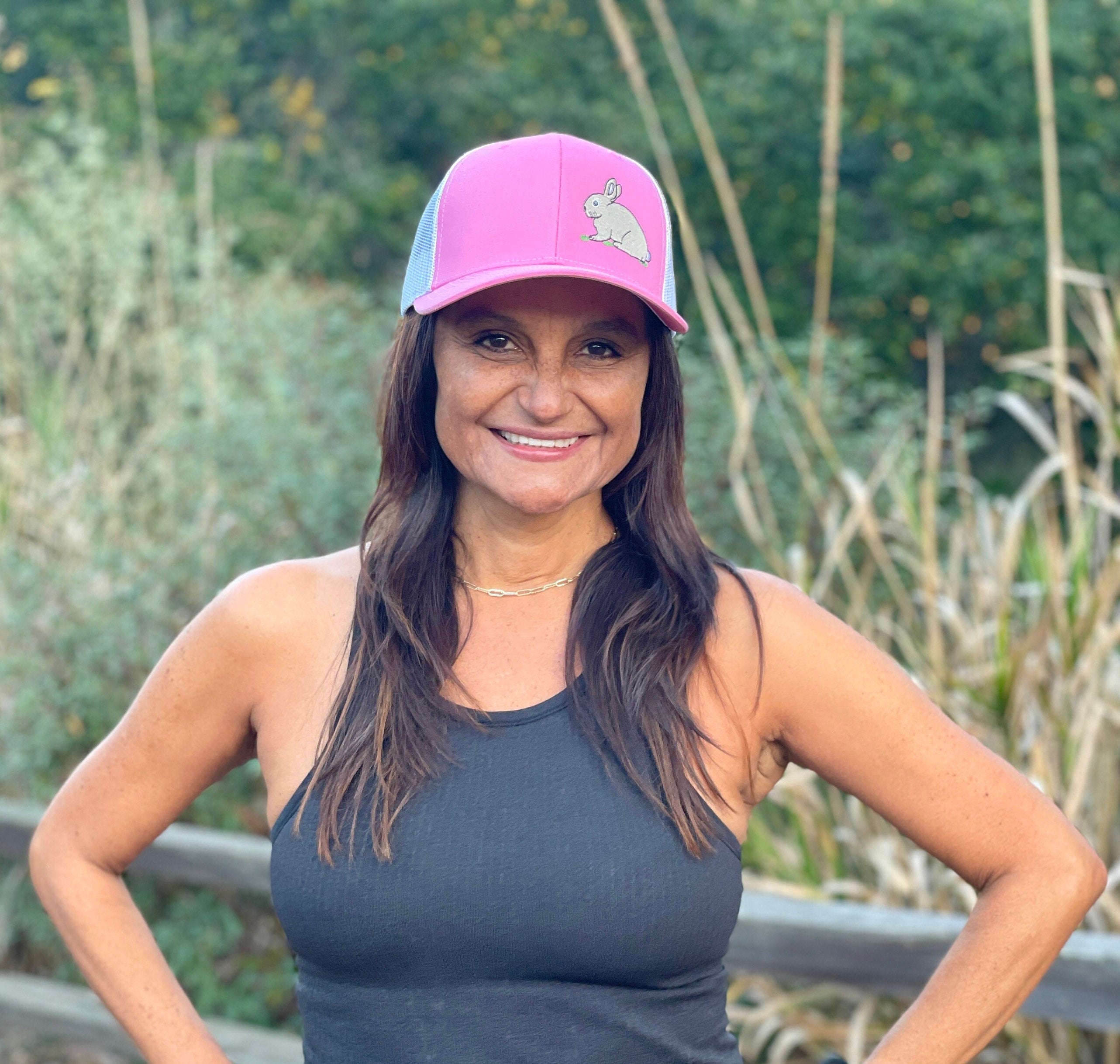 A woman with long dark hair is on a nature trail. She smiles wearing a cute trucker hat that is pink on the front panel with white mesh on the back and a snapback closure. A cute tan bunny in green grass is embroidered on the front of the hat.
