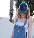 Girl with brown curly hair wears a cute trucker hat that is navy blue on the front with light blue mesh on the back and a velcro closure. Morning glory flowers are embroidered on the front side of the hat.