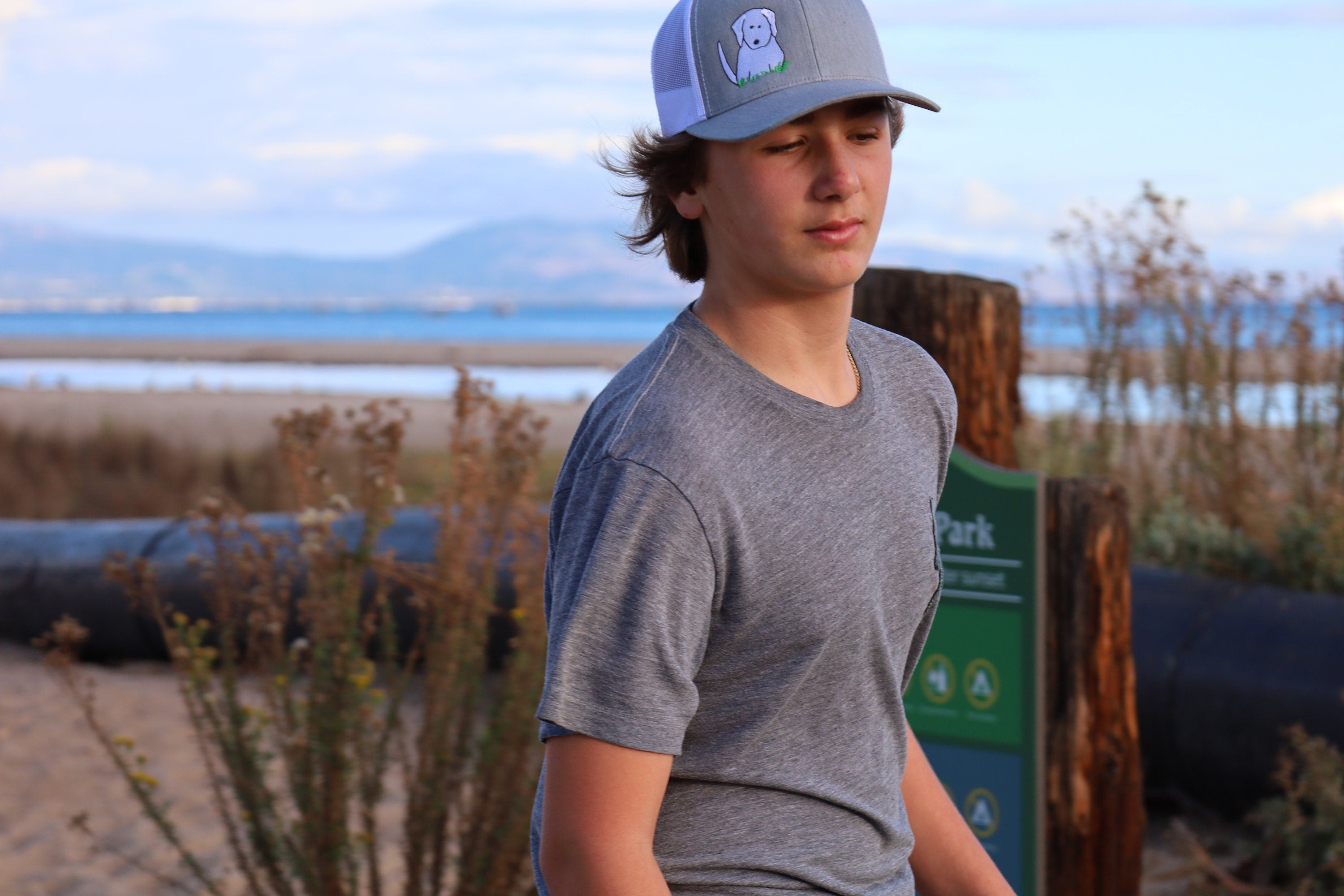 Teenage boy with brown hair stands on the beach with the ocean behind him. He wears a cute trucker hat that is grey on the front panel with white mesh on the back and a snapback closure. A cute white dog is embroidered on the front of the hat.