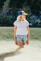 Young woman with blonde hair stands in a river. She wears a vintage inspired trucker hat that is khaki on the front panel with blue mesh on the back and a snapback closure. A mountain scene is embroidered on the front of the hat.