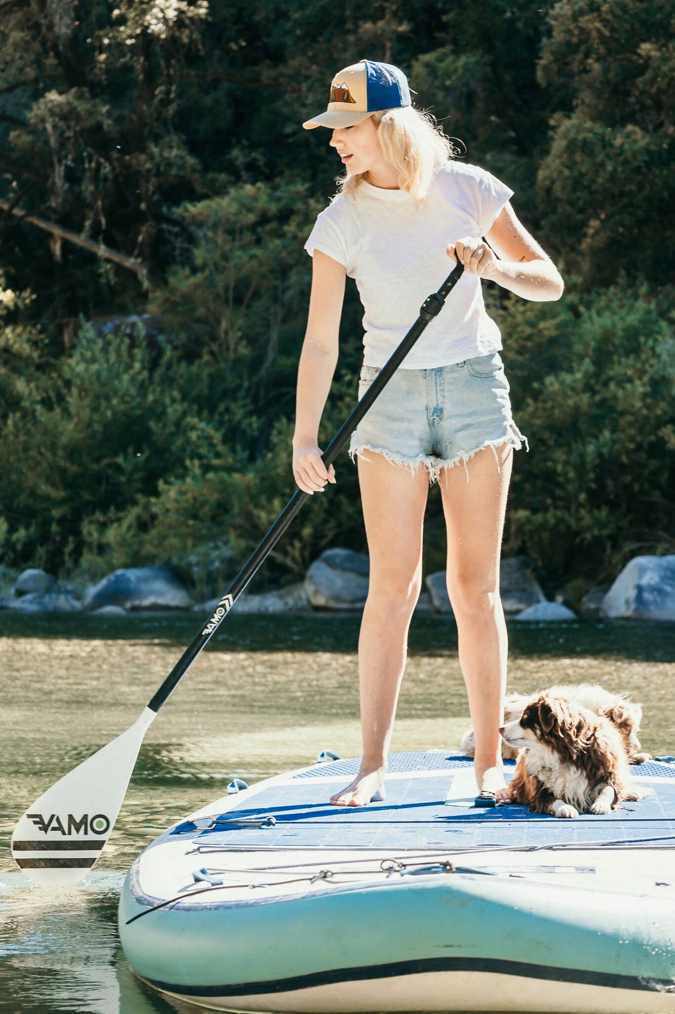 Young woman is on a stand up paddle board with dogs. She wears a vintage inspired trucker hat that is khaki on the front panel with blue mesh on the back and a snapback closure. A mountain scene is embroidered on the front of the hat.