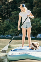 Young woman is on a stand up paddle board with dogs. She wears a vintage inspired trucker hat that is khaki on the front panel with blue mesh on the back and a snapback closure. A mountain scene is embroidered on the front of the hat.