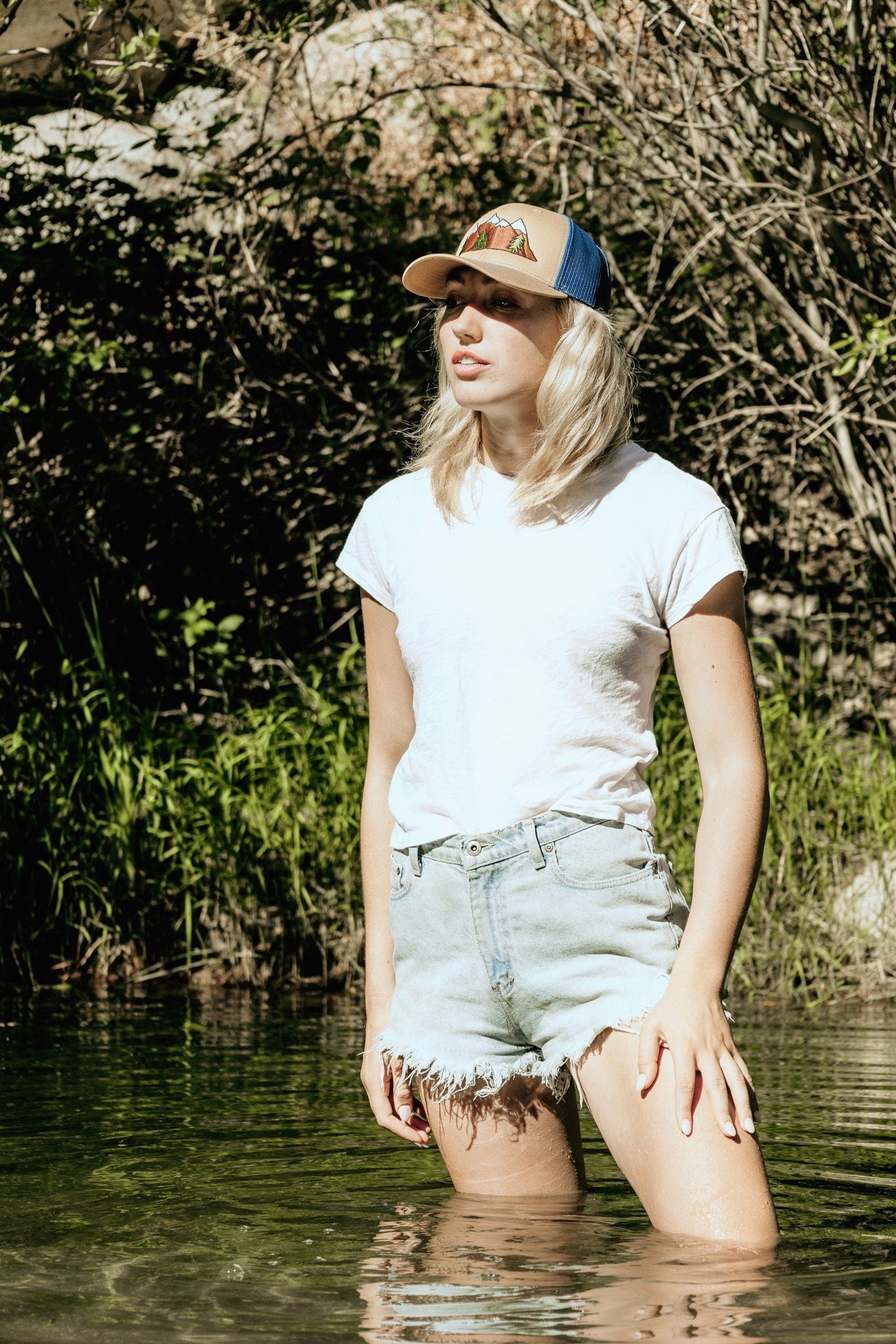 Young woman with blonde hair stands in a river. She wears a cool trucker hat that is khaki on the front panel with blue mesh on the back and a snapback closure. A mountain scene is embroidered on the front of the hat.
