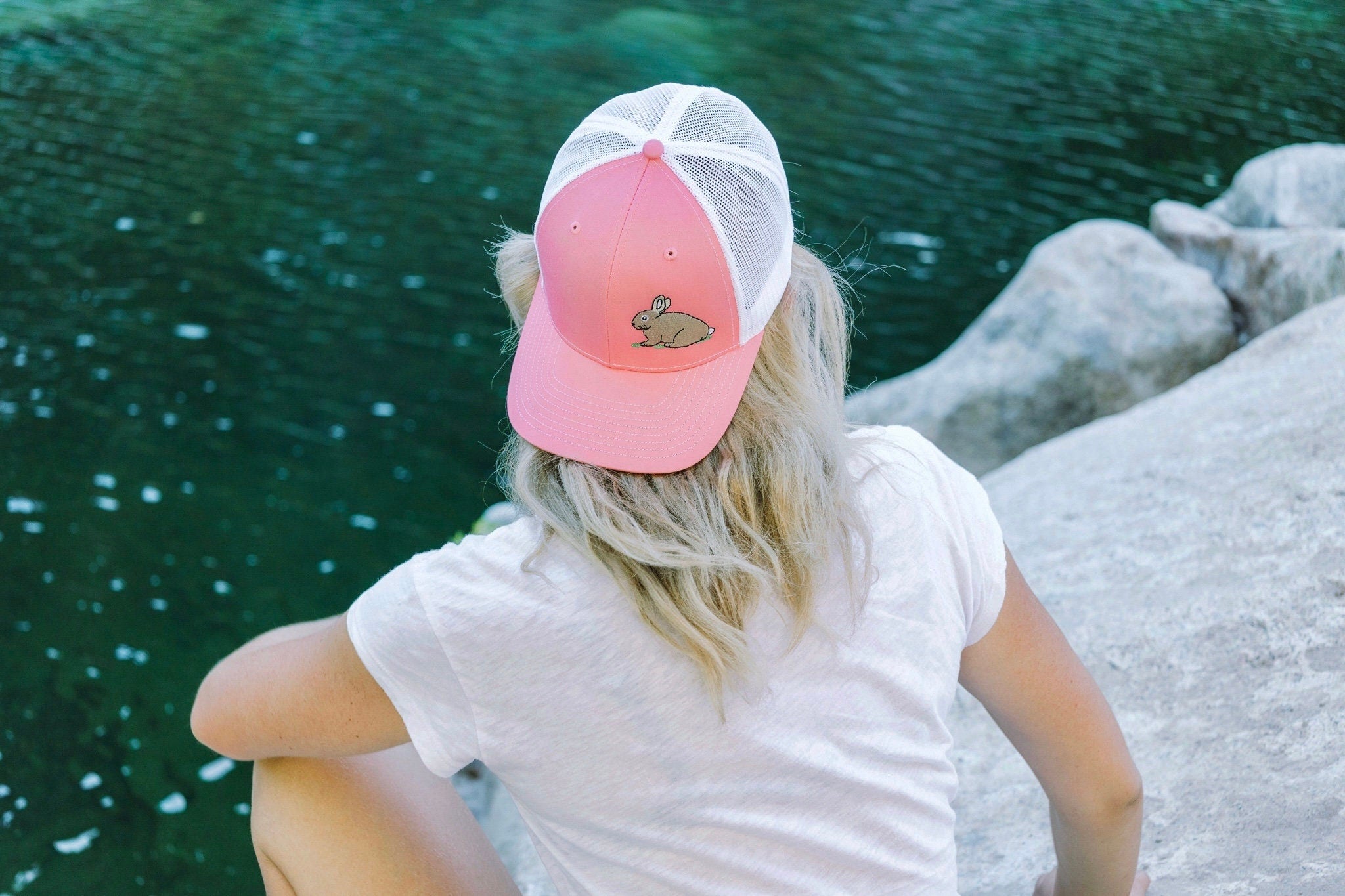 Young woman with blonde hair sits facing a river. She wears a cute trucker hat backwards that is pink on the front panel with white mesh on the back and a snapback closure. A cute tan bunny in green grass is embroidered on the front of the hat.