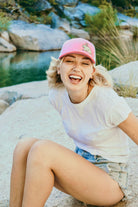 Young woman with blonde hair sits on a rock by a river. She wears a cute trucker hat that is pink on the front panel with white mesh on the back and a snapback closure. A cute tan bunny in green grass is embroidered on the front of the hat.
