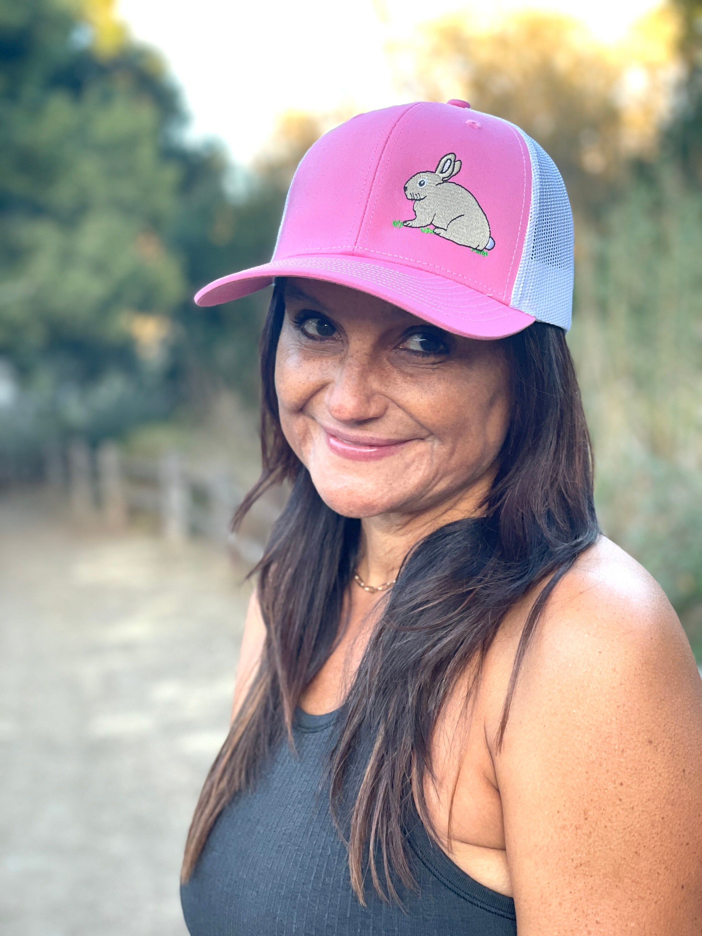 A woman with long dark hair is on a nature trail. She smiles wearing a cute trucker hat that is pink on the front panel with white mesh on the back and a snapback closure. A cute tan bunny in green grass is embroidered on the front of the hat.
